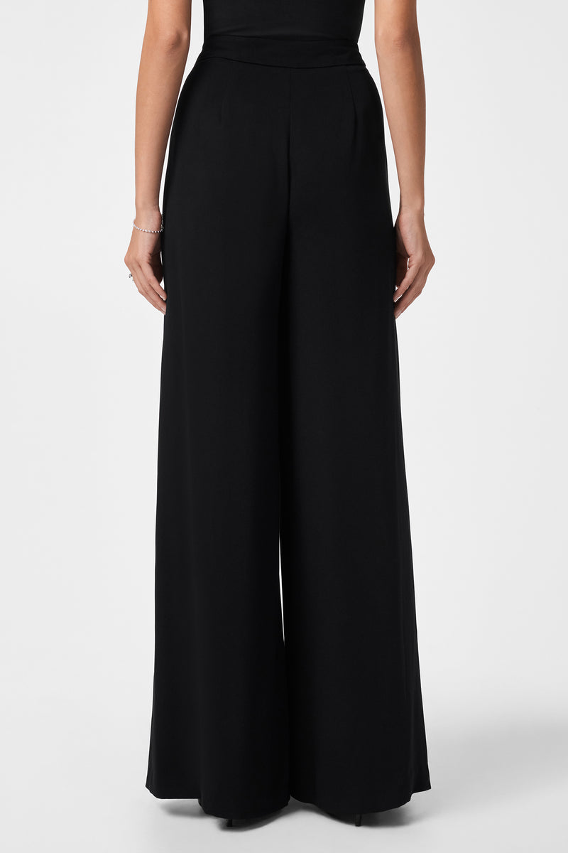 A person wearing the Saige Long Pant—high-waisted, wide-leg black pants—and a black top is shown from the back against a plain white background.
