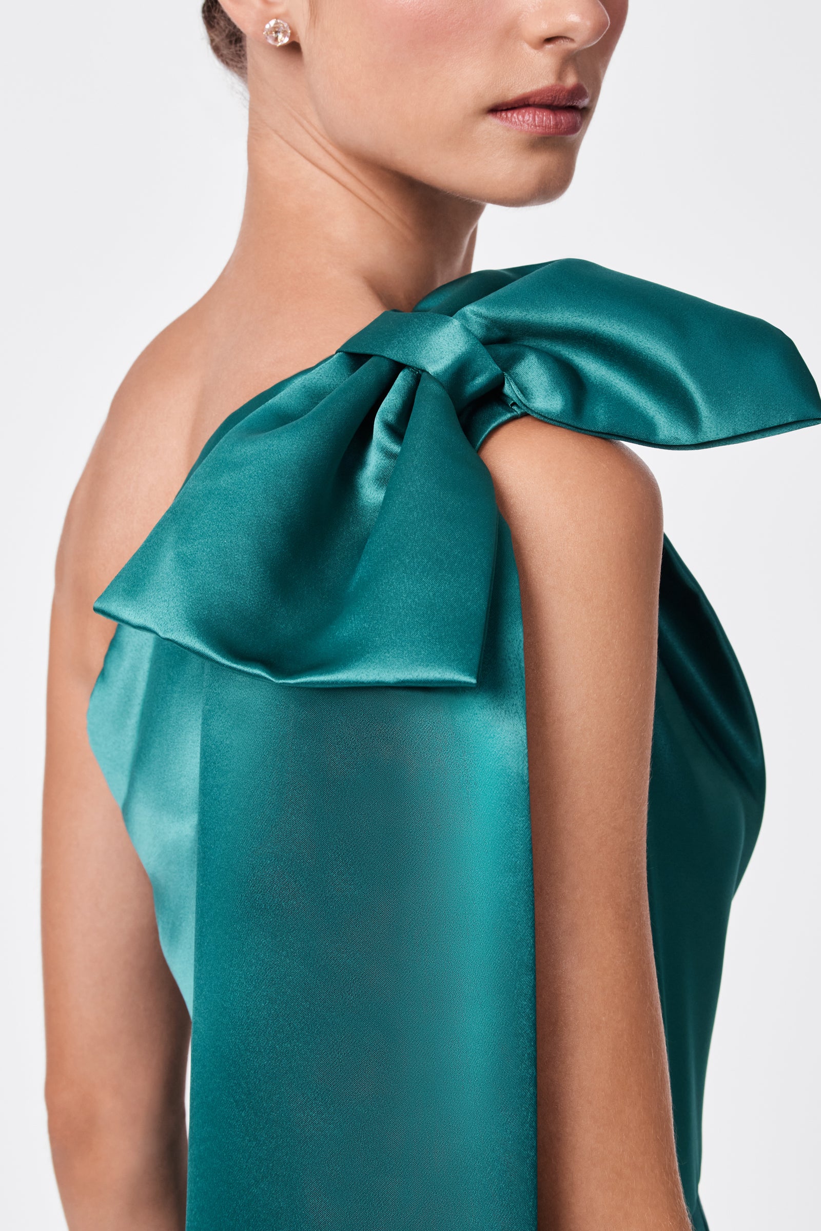 A woman with light skin wears the teal satin Romance Dress, featuring a large, elegant bow on one shoulder. She is shown from the lips down, highlighting the dress detail and her small stud earring.