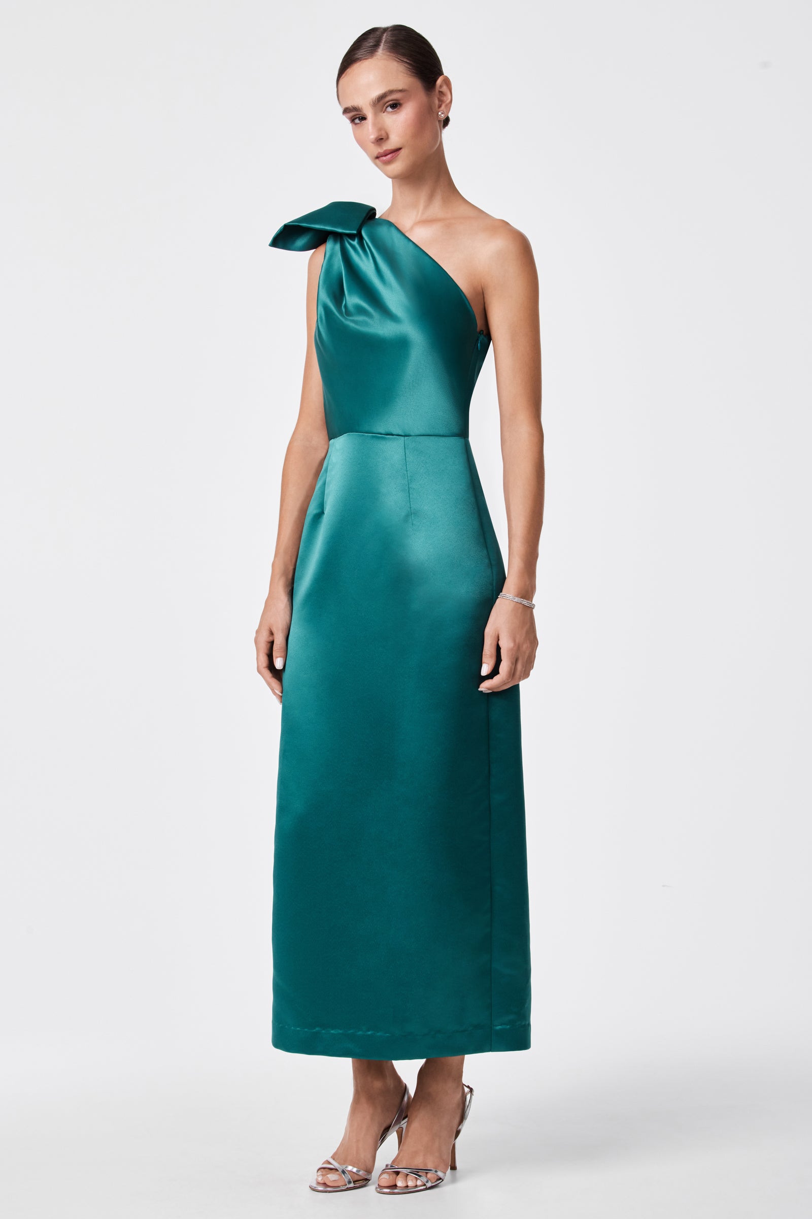 A woman stands facing forward in the Romance Dress—a teal satin, one-shoulder design with a bow detail—her hair pulled back and paired with silver high-heeled sandals, set against a plain white background.