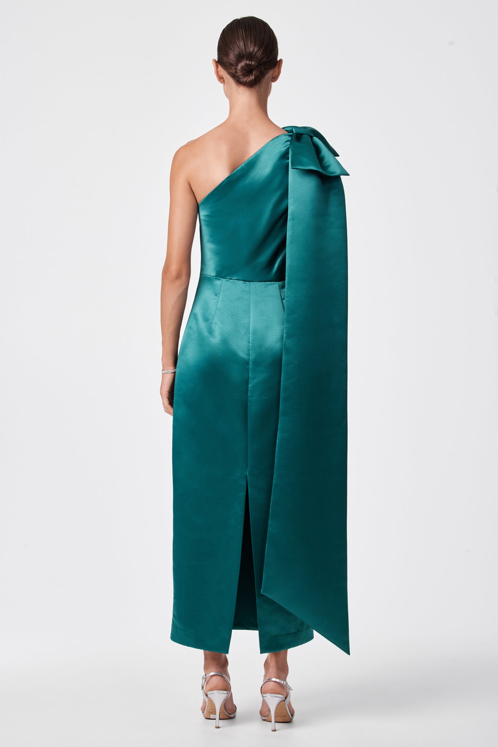 A woman, hair in a low bun, models the Romance Dress—a teal one-shoulder satin gown featuring a large bow with a long drape detail. She pairs it with silver high-heeled sandals and faces away from the camera.