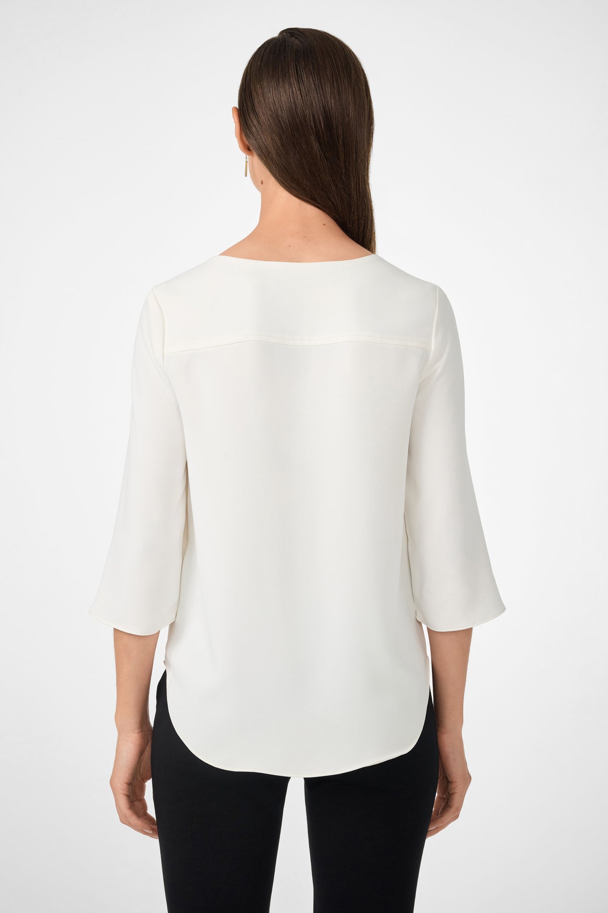 A woman with straight brown hair, seen from the back, wears the white Tamarac Top with three-quarter sleeves and black pants against a plain white background.