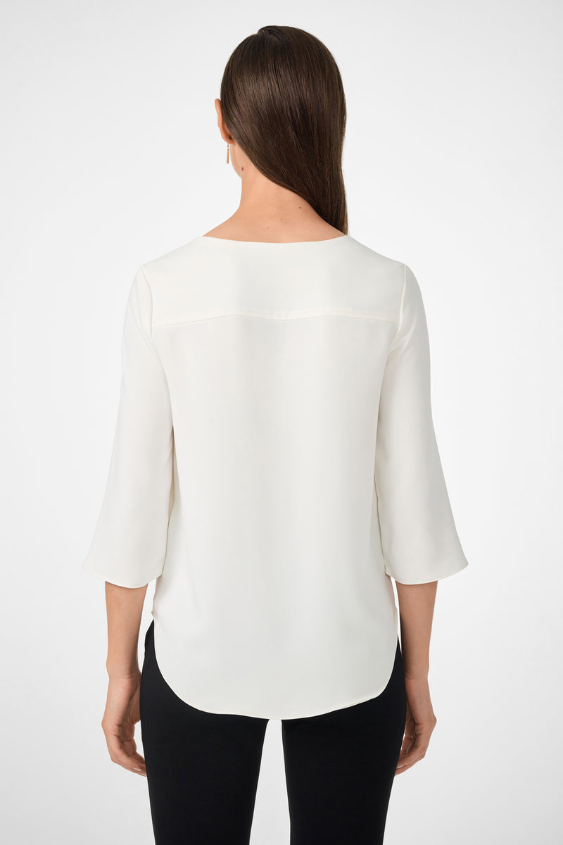 A woman with straight brown hair, seen from the back, wears the white Tamarac Top with three-quarter sleeves and black pants against a plain white background.