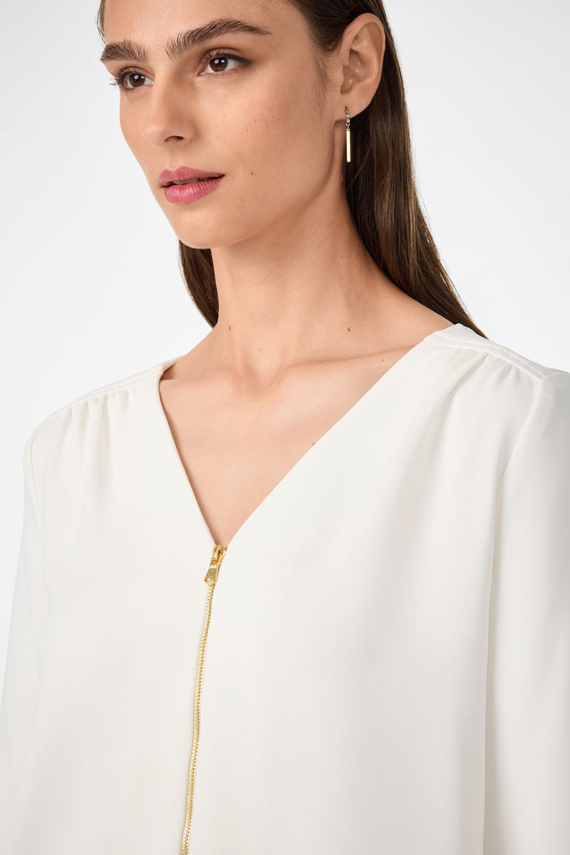 A woman with straight brown hair wears the Tamarac Top, a white V-neck blouse with a gold zipper, and a gold hoop earring. The photo is from the shoulders up against a plain light background.