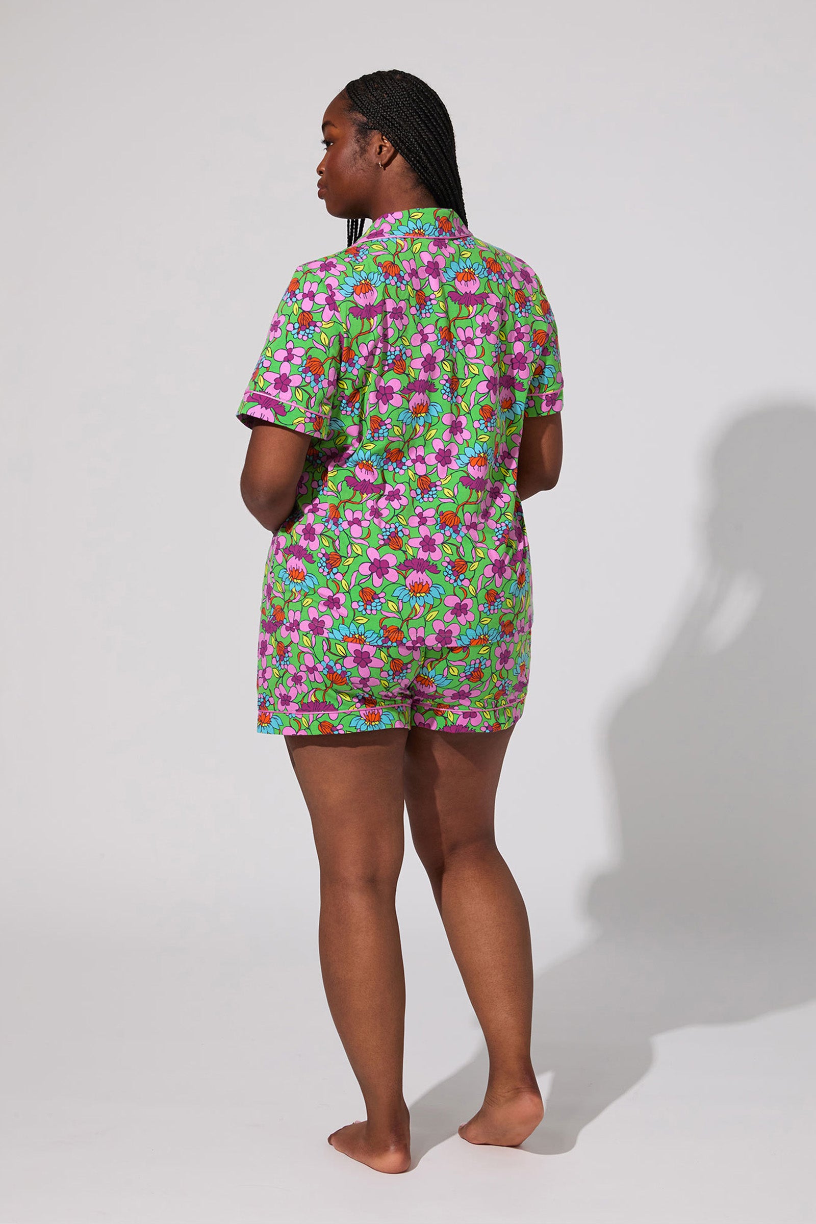A person stands barefoot facing away, wearing the Shorty PJ Set - Garden Floral, a matching short-sleeve shirt and shorts with a green, pink, and purple floral print. The background is plain and light-colored.