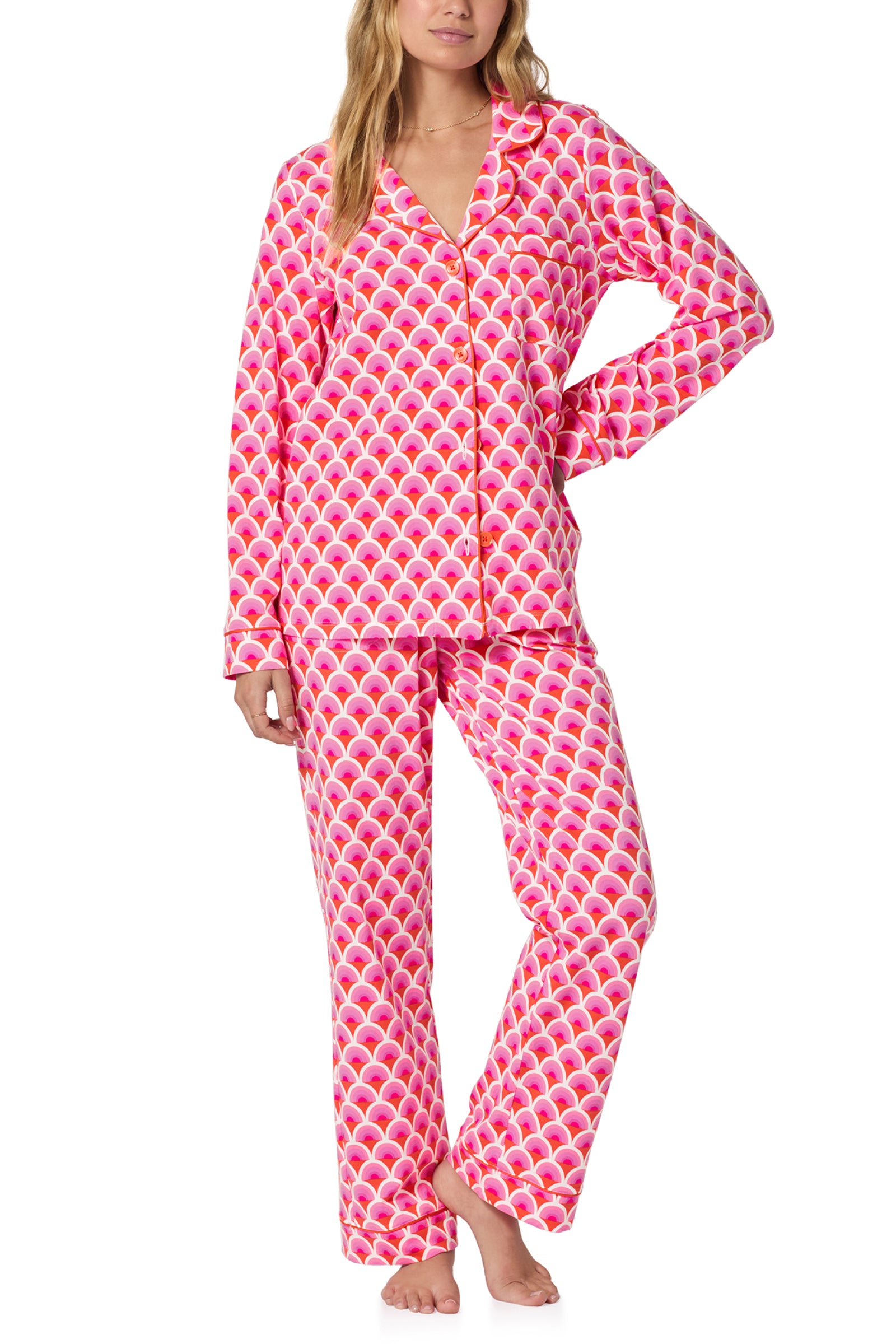 A woman stands barefoot in the Classic PJ Set - Scallop, featuring a matching pink and red scallop pattern with a long-sleeve button-up top and loose-fitting pants.