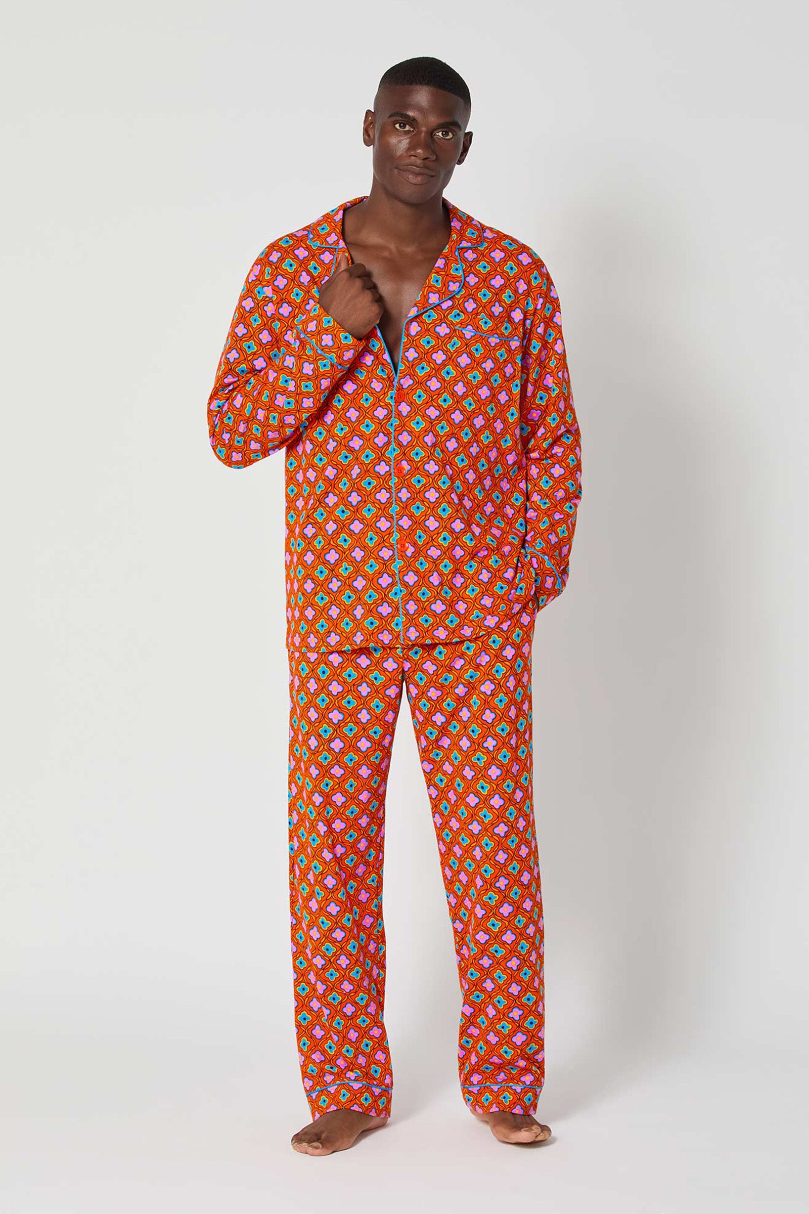 A man stands barefoot against a plain background, wearing the Daisy Foulard Mens Classic Pj—a matching red pajama set with a colorful geometric pattern—holding the edge of the open top with one hand on his chest.