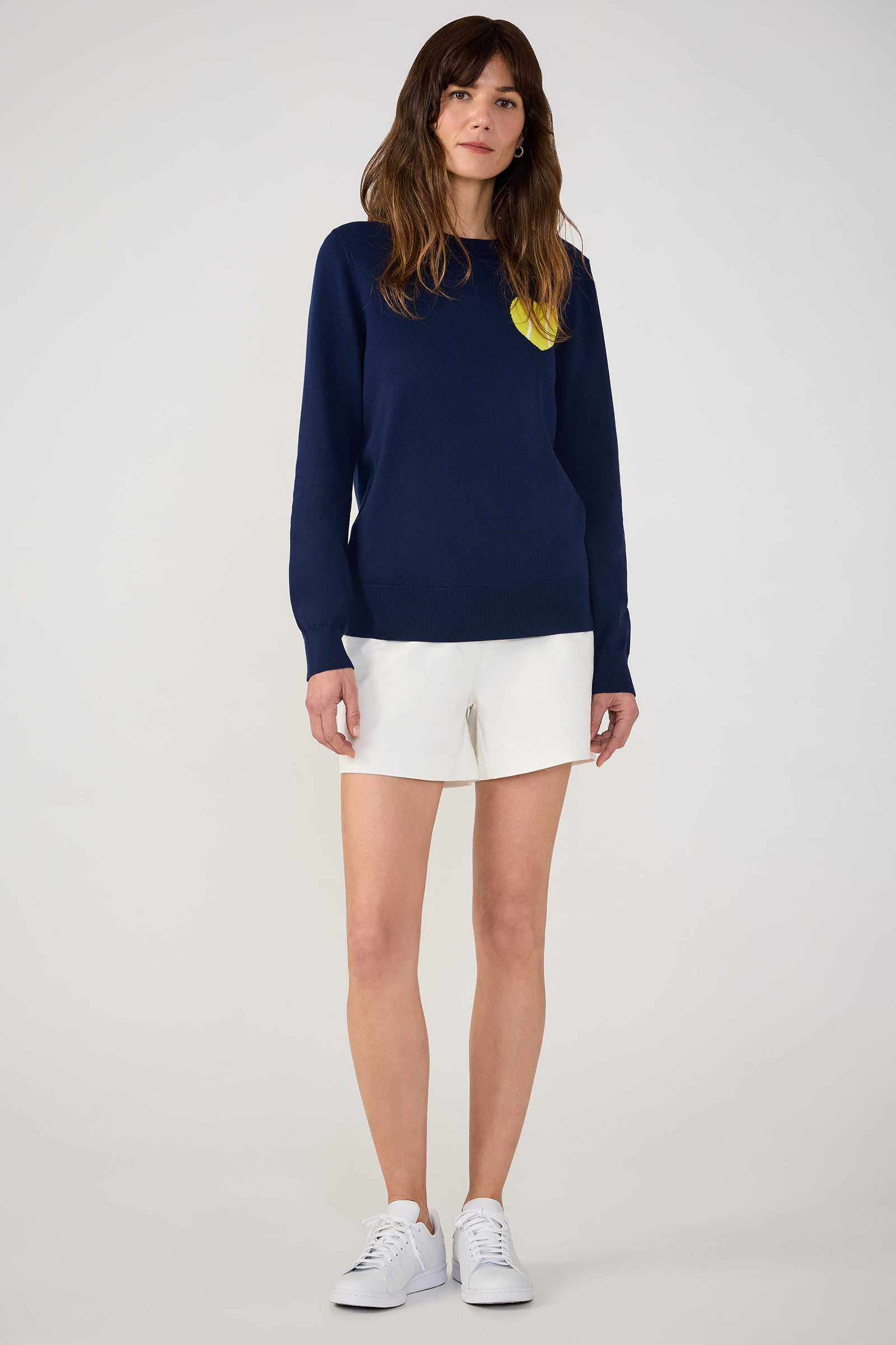 A woman faces forward against a plain light background, wearing the Tennis Heart Long-Sleeve Crewneck Sweater in navy blue with a yellow chest accent, paired with white shorts and white sneakers.