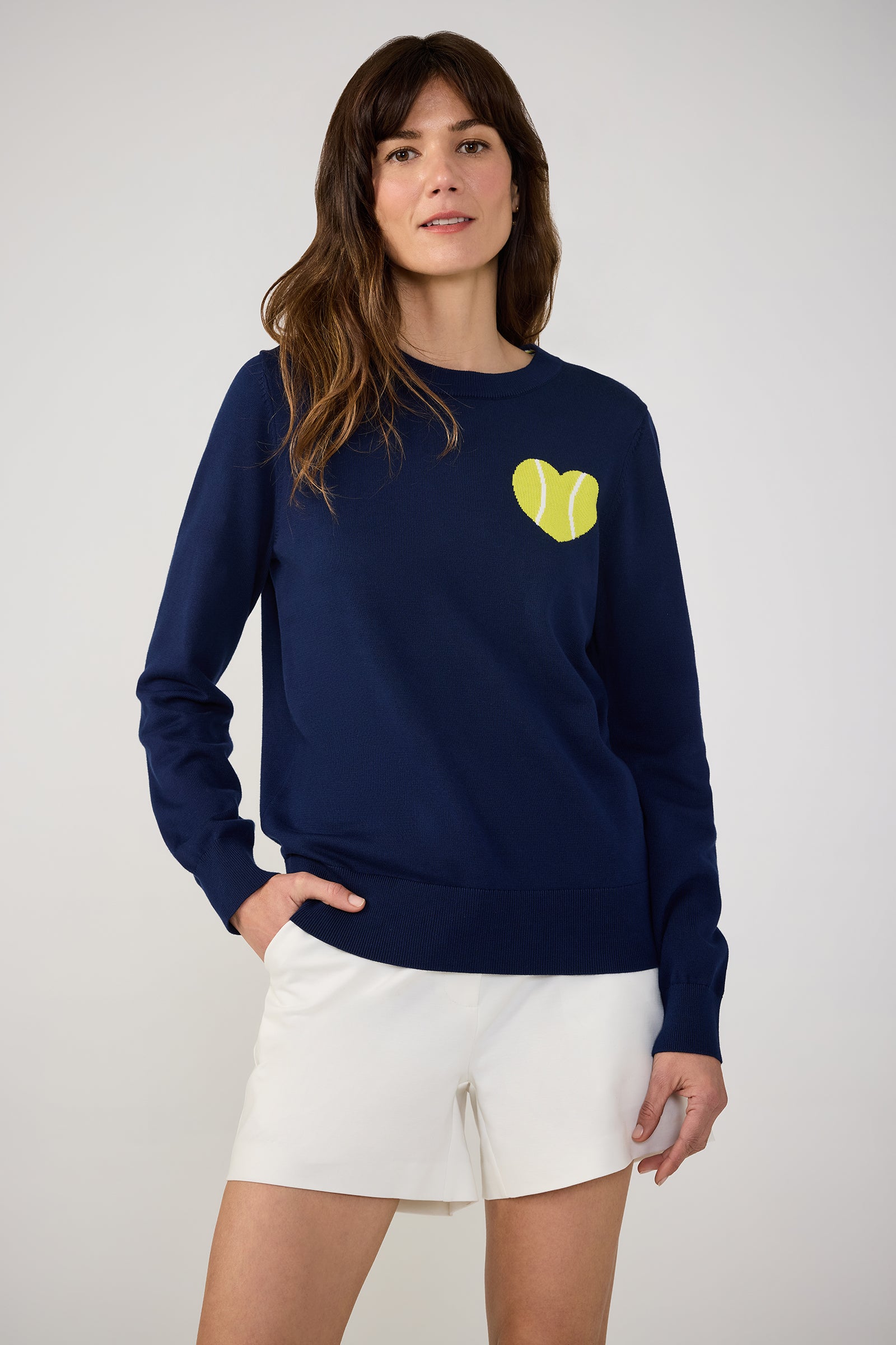 A woman with long brown hair wears the Tennis Heart Long-Sleeve Crewneck Sweater in Ink, featuring a yellow tennis ball graphic on the chest, paired with white shorts and standing against a plain light background.