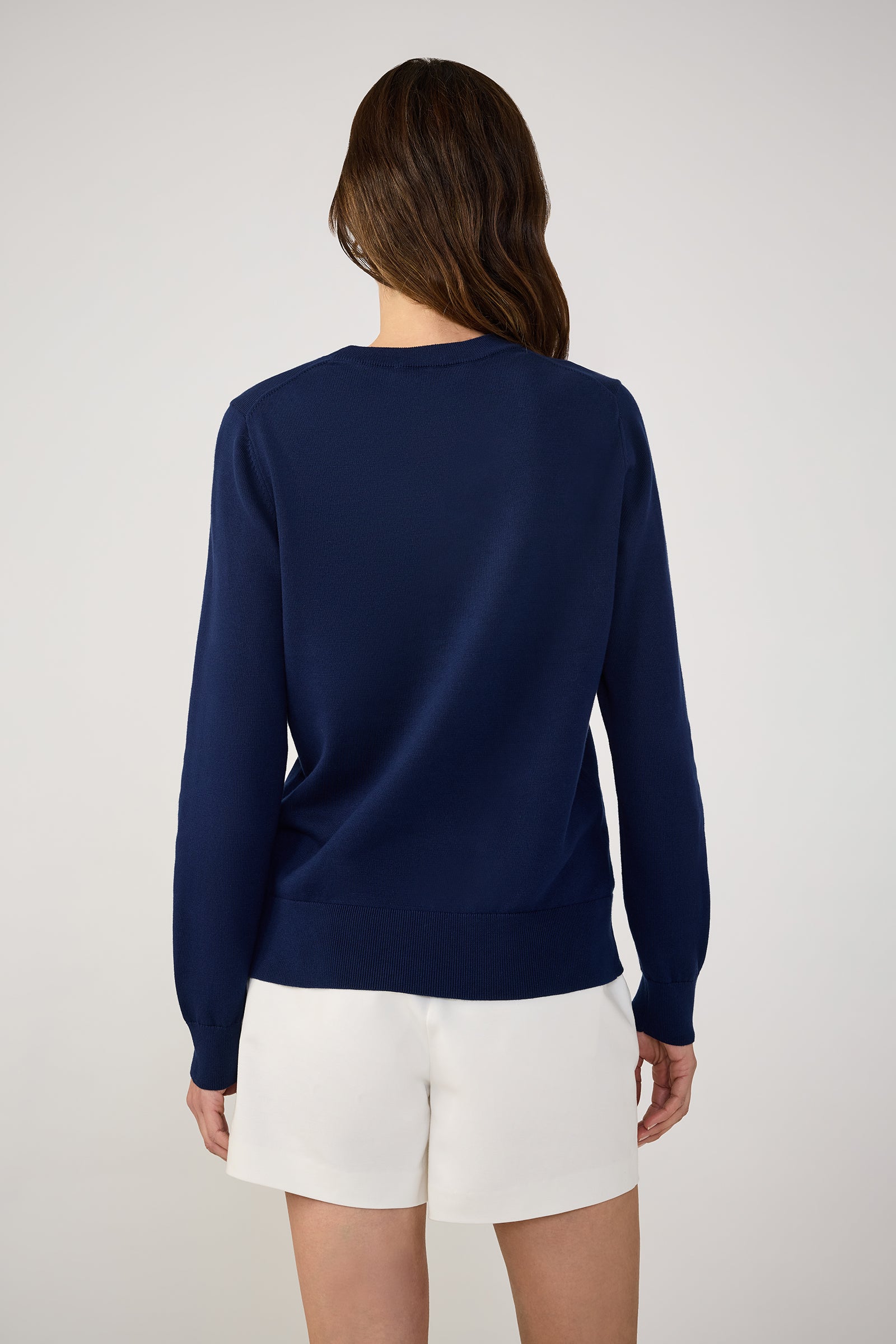 A person with long brown hair is shown from the back, wearing the Tennis Heart Long-Sleeve Crewneck Sweater in navy blue and white shorts, standing against a plain light background.