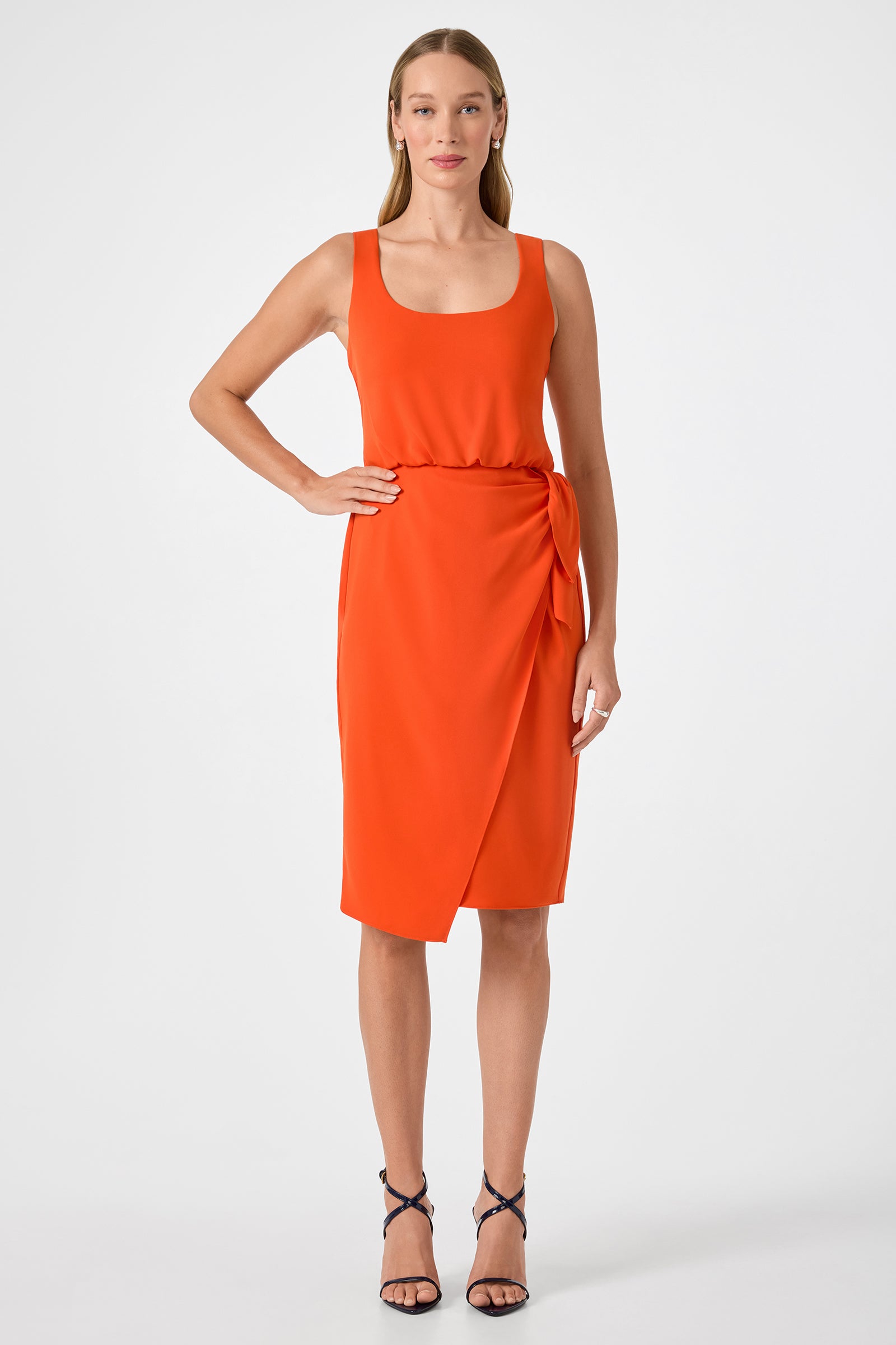 A woman stands against a white background wearing the ORita Sleeveless Tie-Waist Midi Dress in bright orange, featuring a wrap-style skirt and side tie, paired with black strappy heels.