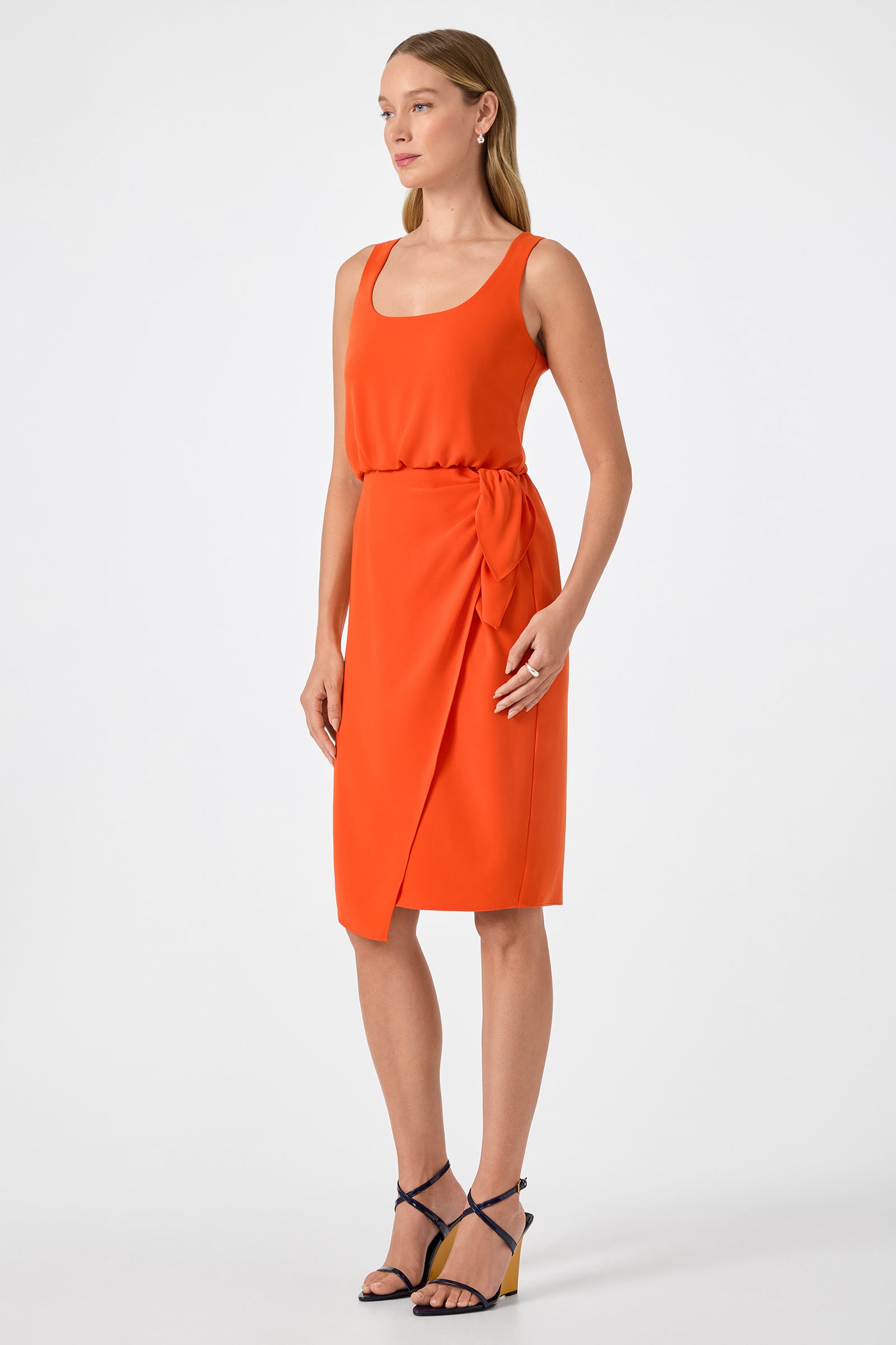 A woman models the ORita Sleeveless Tie-Waist Midi Dress, a bright orange, knee-length dress with a wrapped skirt and waist tie. She pairs it with black strappy heels against a plain white background.