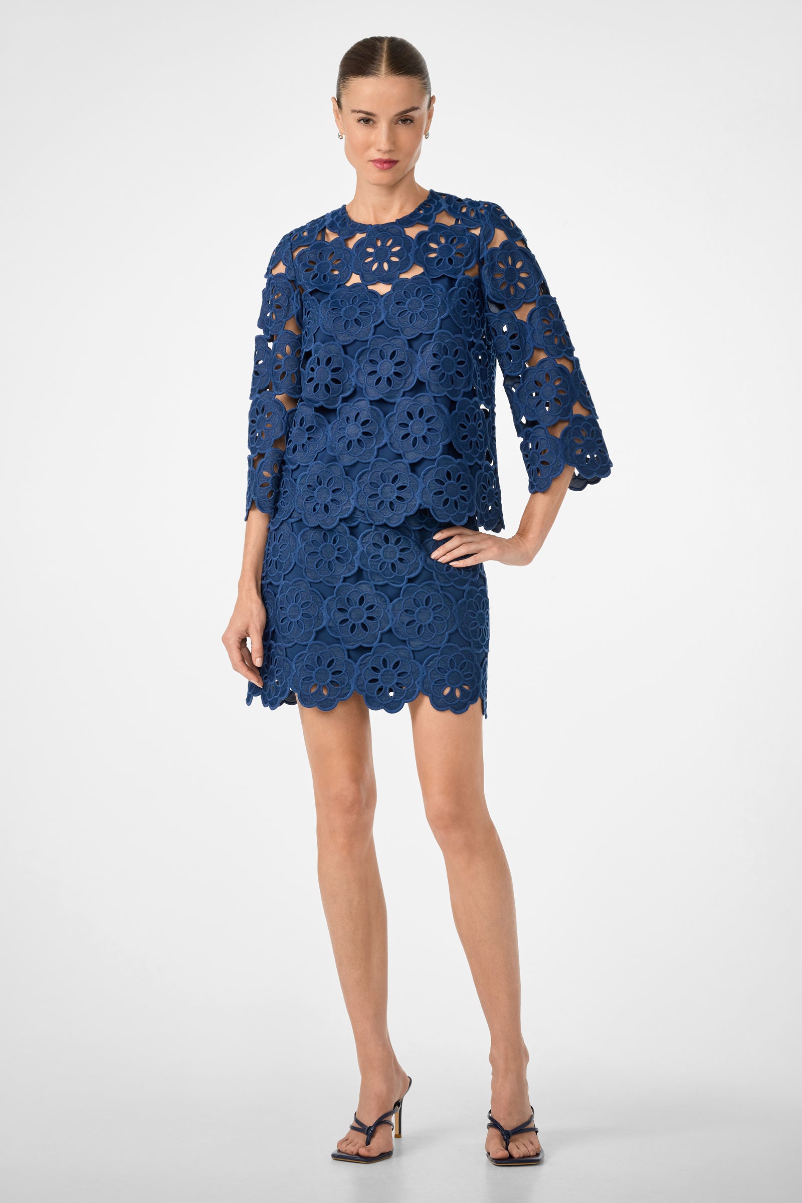 Woman wearing the High Rise Rico Skirt in Kusho Embroidery, featuring bold floral patterns and a knee-length silhouette. She stands confidently in strappy black heels against a plain backdrop.