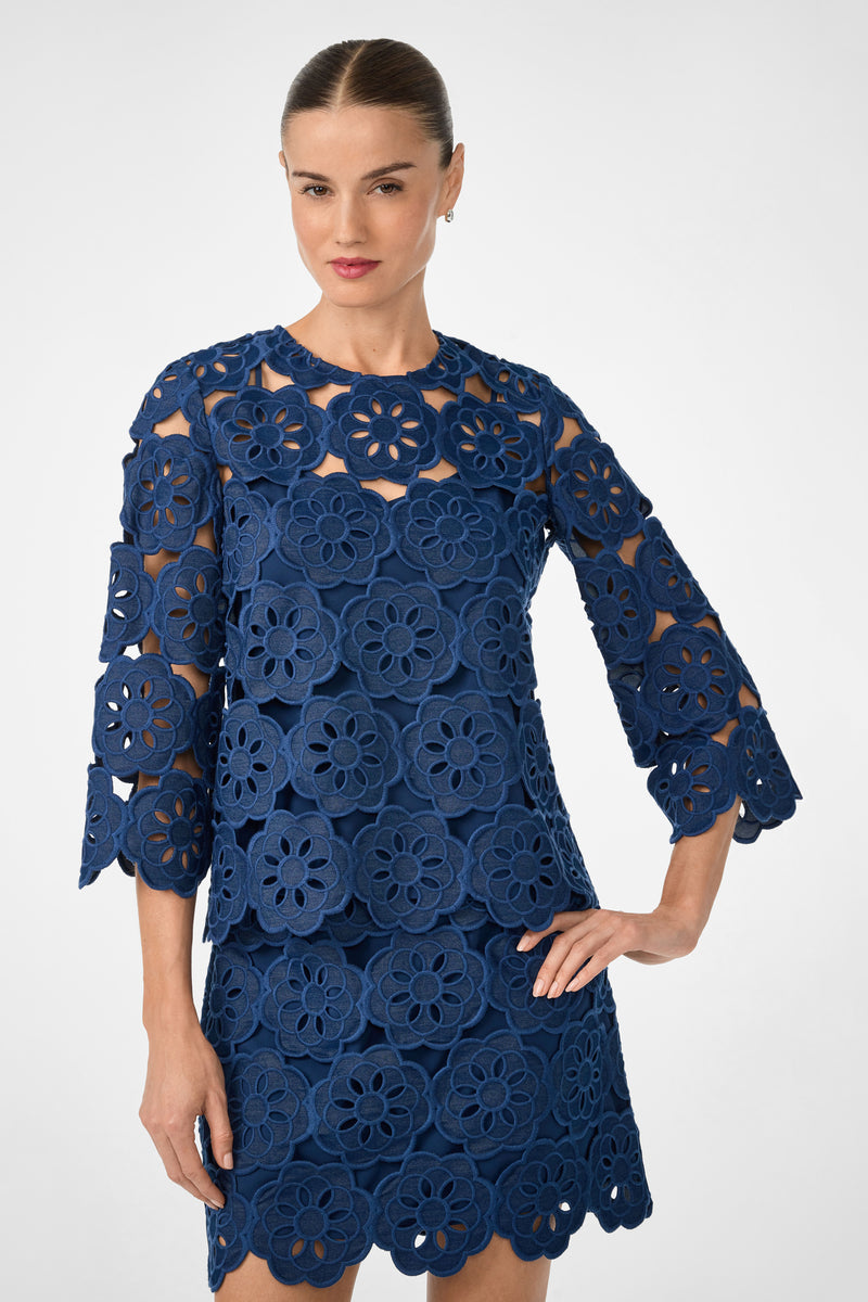 A woman wears the Kabosu Top - Kusho Embroidery in indigo, featuring bold floral cutout patterns, three-quarter sleeves, and a round neckline. Her hair is slicked back as she poses against a plain light background.