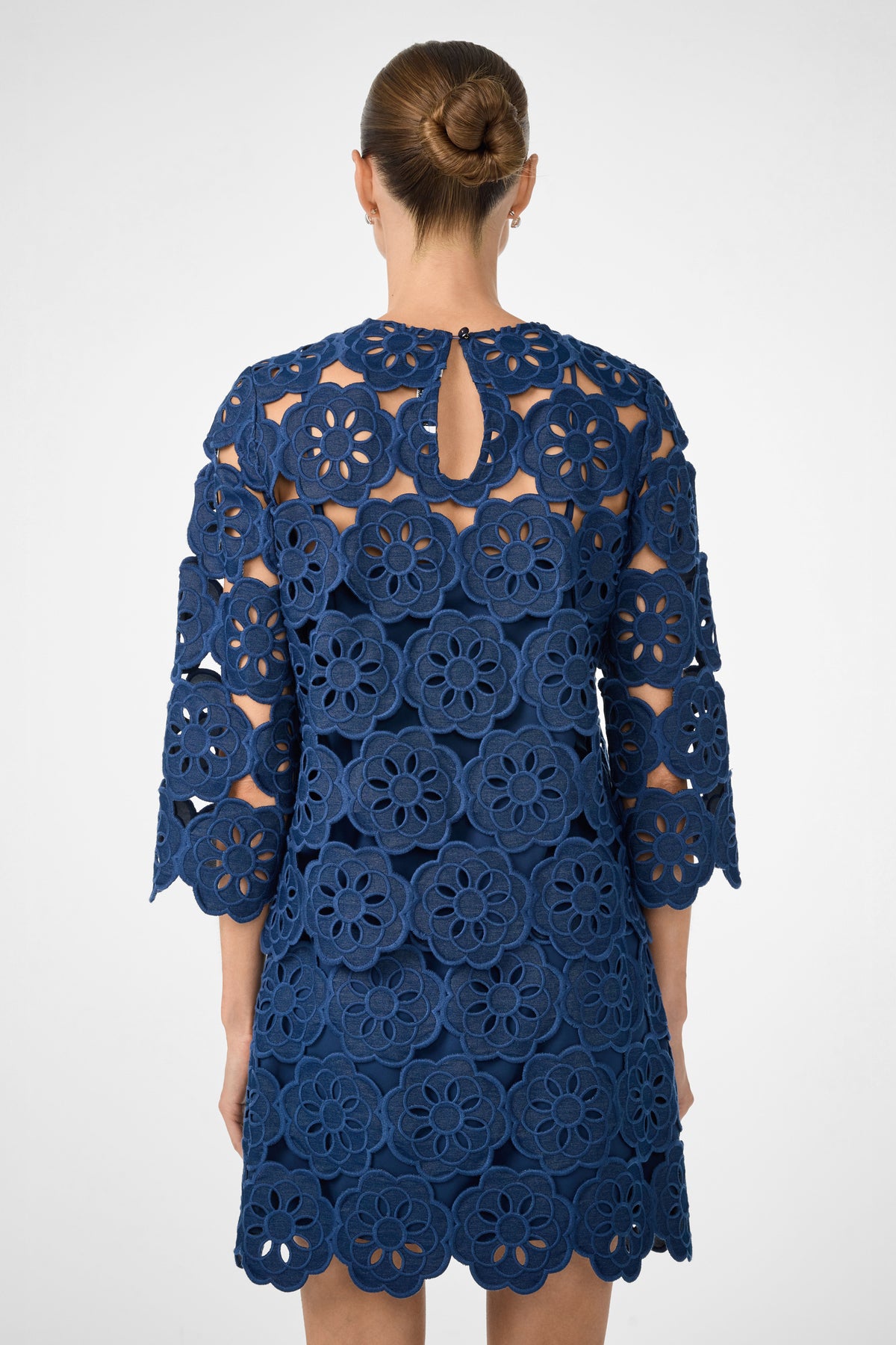 A woman with her hair in a bun is seen from the back wearing the Kabosu Top - Kusho Embroidery, a navy blue piece featuring large floral cutout lace patterns and three-quarter sleeves.