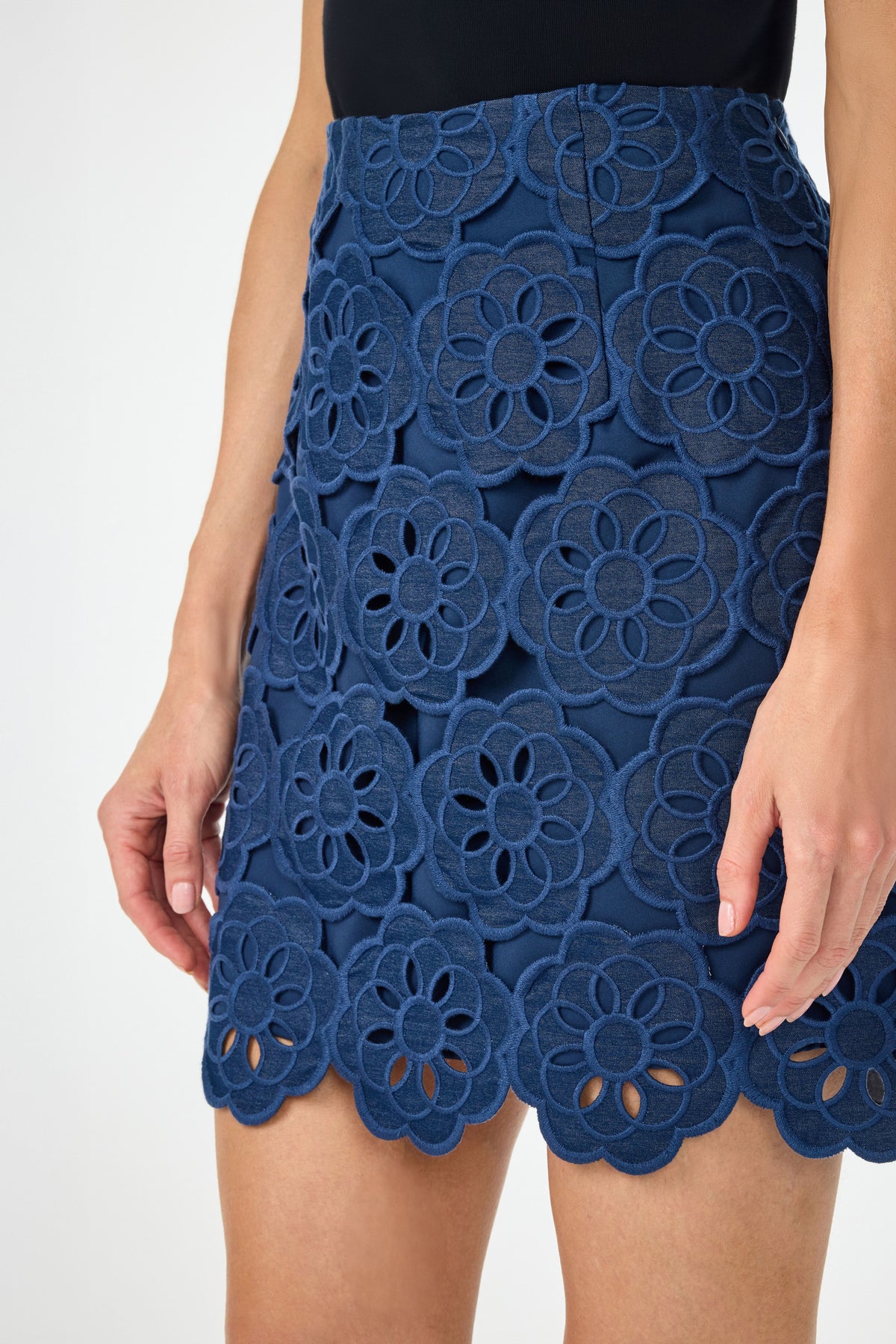 A person wears the High Rise Rico Skirt - Kusho Embroidery, featuring a navy hue with large floral lace patterns and scalloped edges. The black sleeveless top complements the skirt. Only the lower torso and hands show against a plain backdrop.