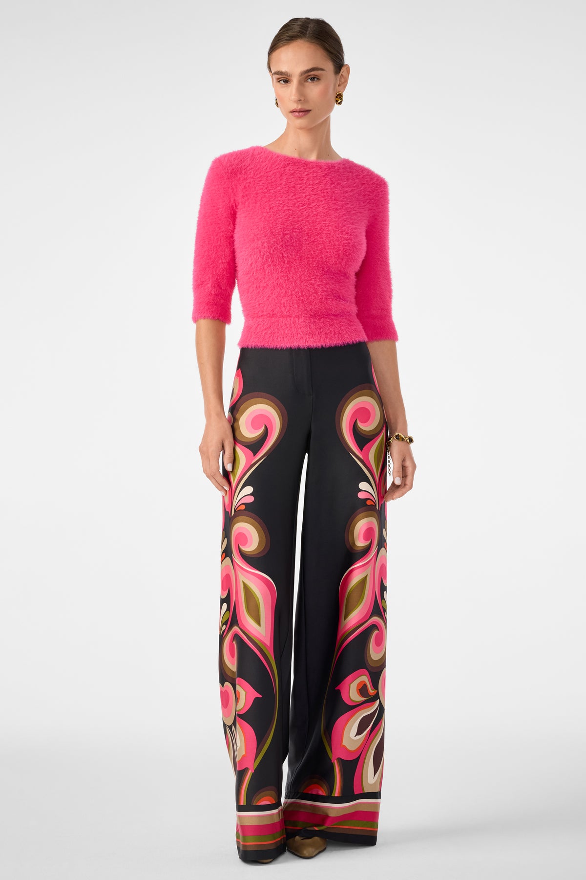 A woman wears the Maliah Fuzzy Sweater in bright pink and high-waisted, wide-leg black pants with a bold swirling pink, red, and beige pattern. Her hair is pulled back and she finishes the look with brown shoes.
