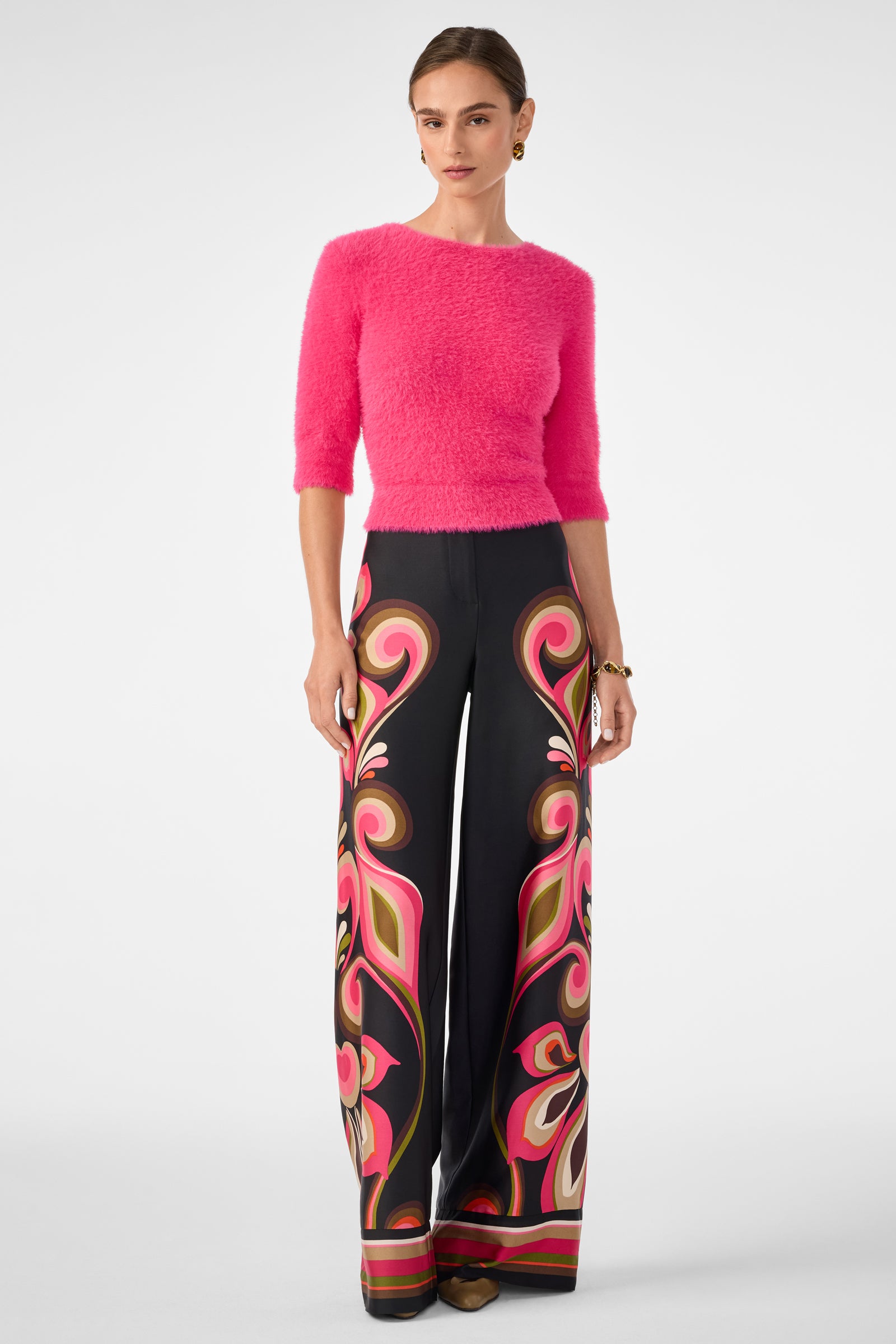 A woman wears the Maliah Fuzzy Sweater in bright pink and high-waisted, wide-leg black pants with a bold swirling pink, red, and beige pattern. Her hair is pulled back and she finishes the look with brown shoes.