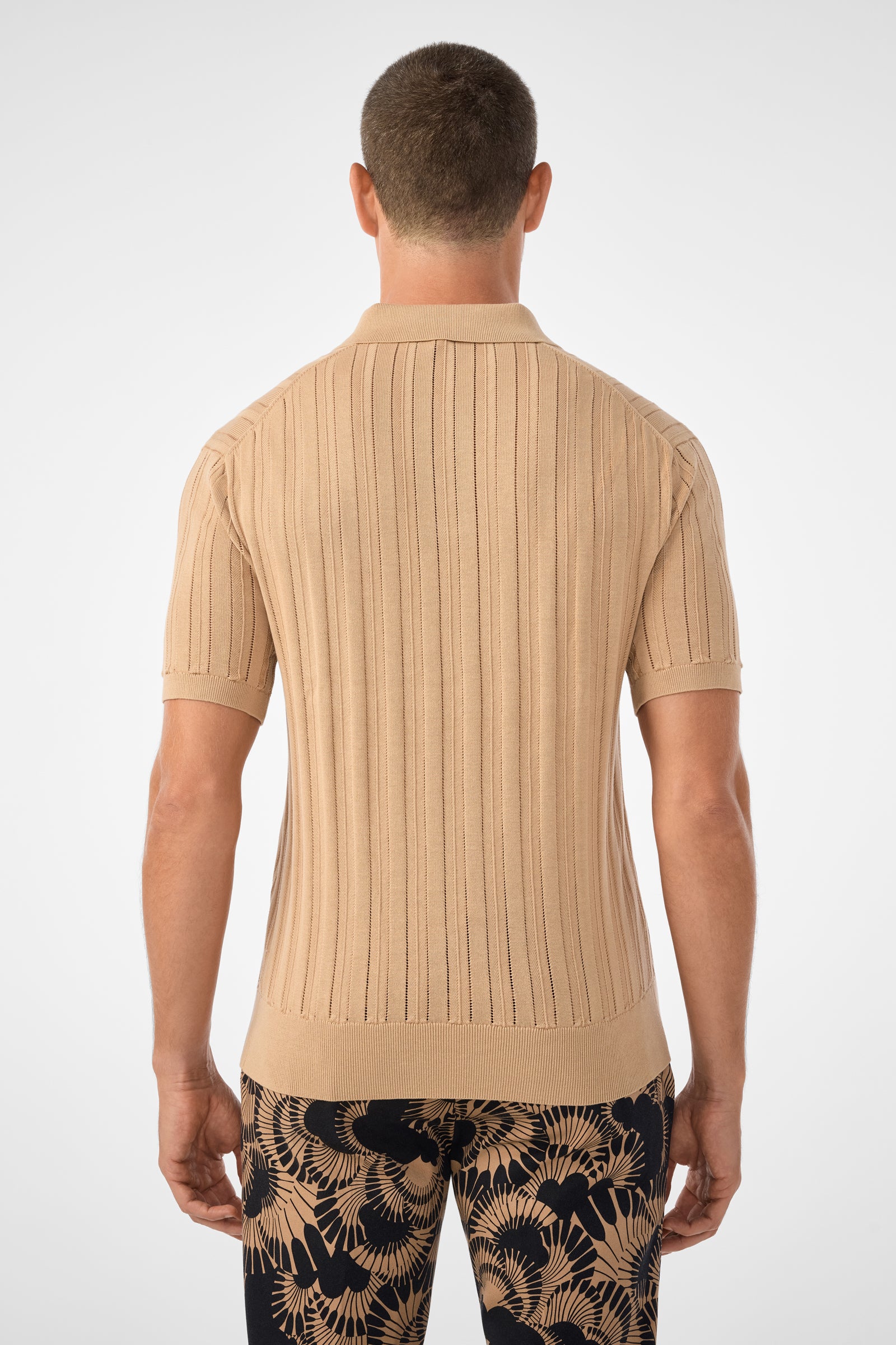 A man stands with his back to the camera, wearing the Kensington Short-Sleeve Polo in beige and navy pants with tan botanical patterns, set against a plain light background.
