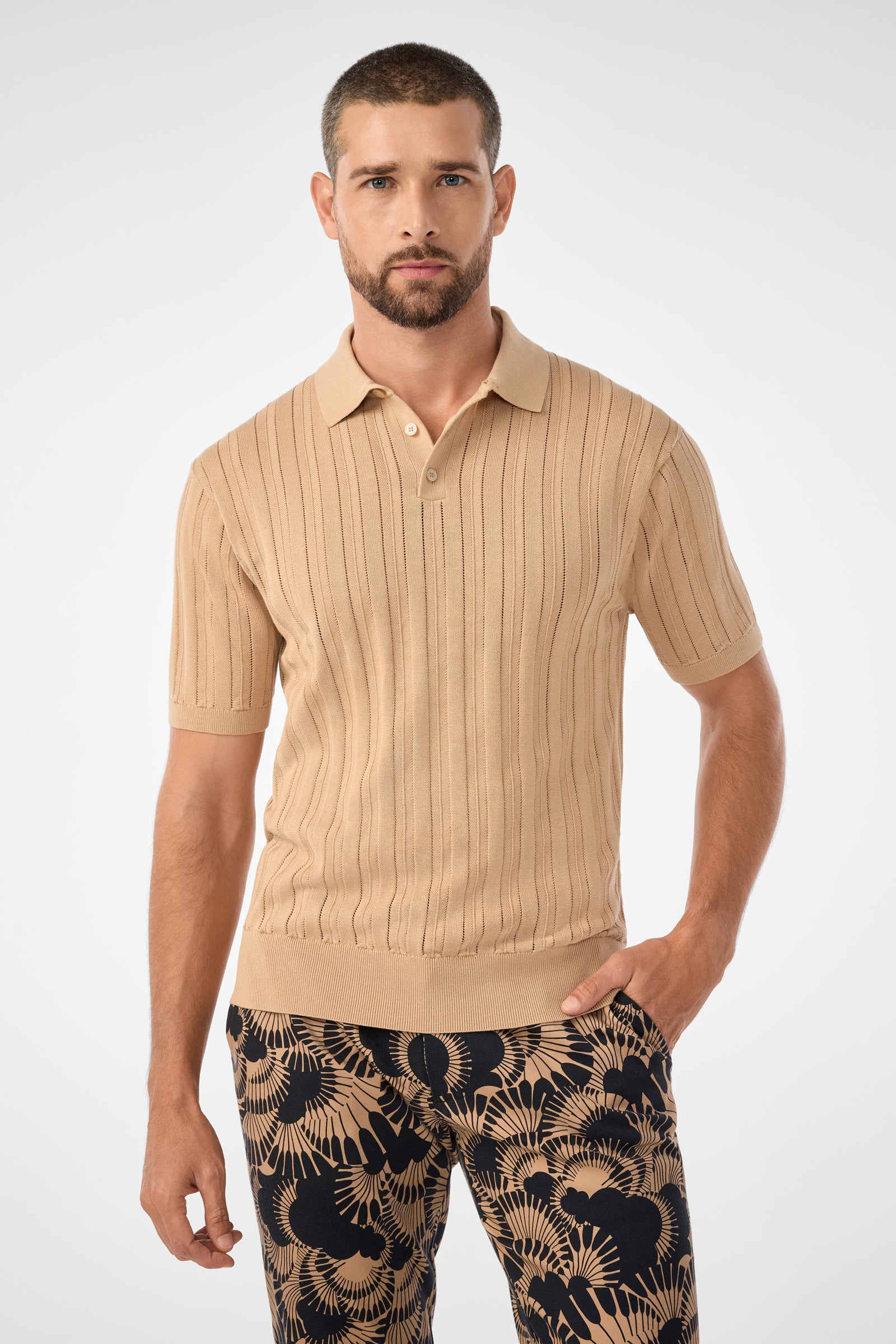 A man with short hair and a beard wears the camel Kensington Short-Sleeve Polo and patterned black pants with tan botanical designs, standing against a plain light gray background.
