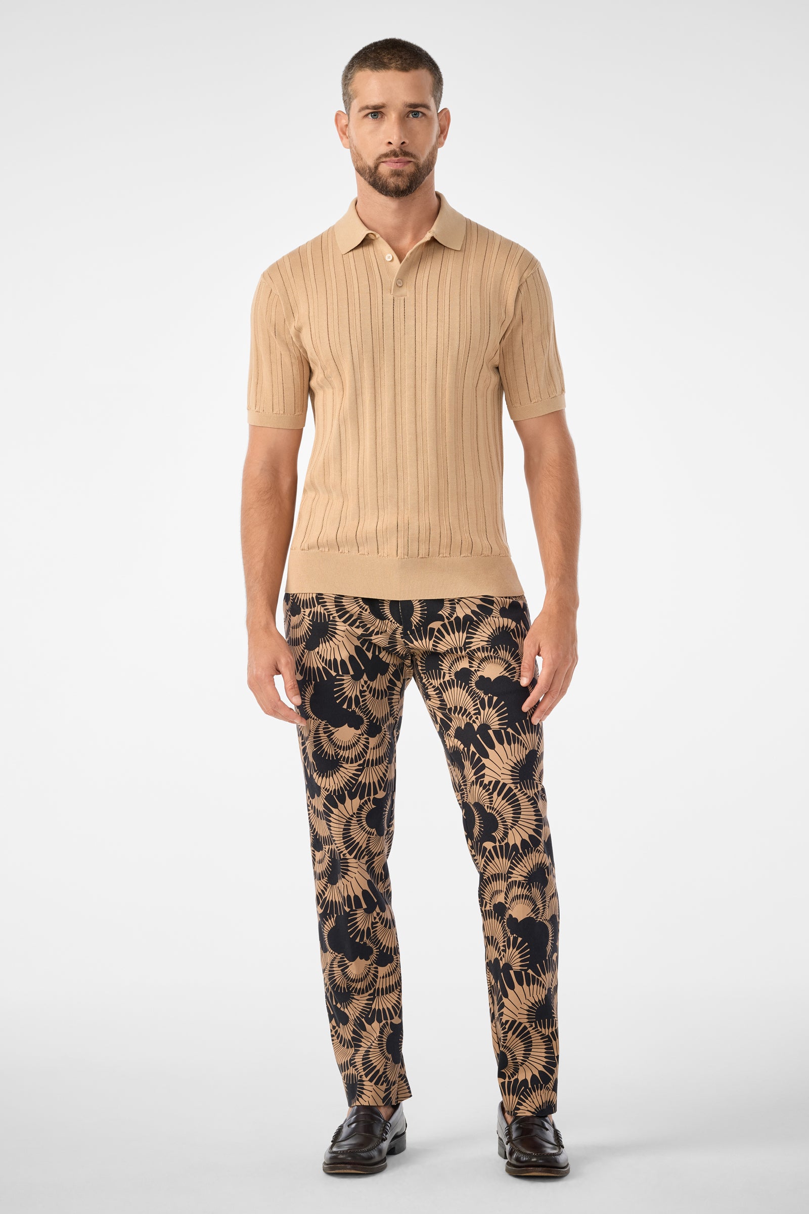 A man stands facing forward against a plain white background, wearing the Kensington Short-Sleeve Polo in beige and black pants with an abstract beige leaf pattern, paired with black dress shoes.