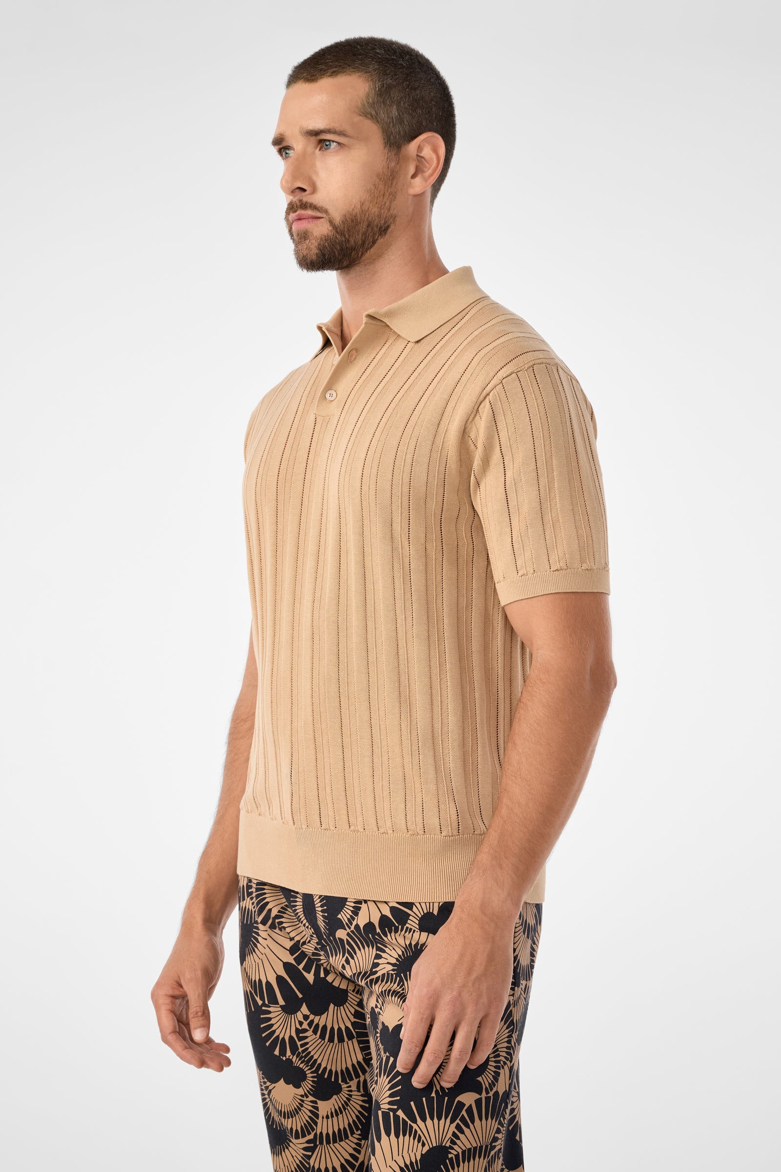 A man with short hair and a beard wears the beige Kensington Short-Sleeve Polo and black pants with a tan botanical pattern, standing against a plain light background.