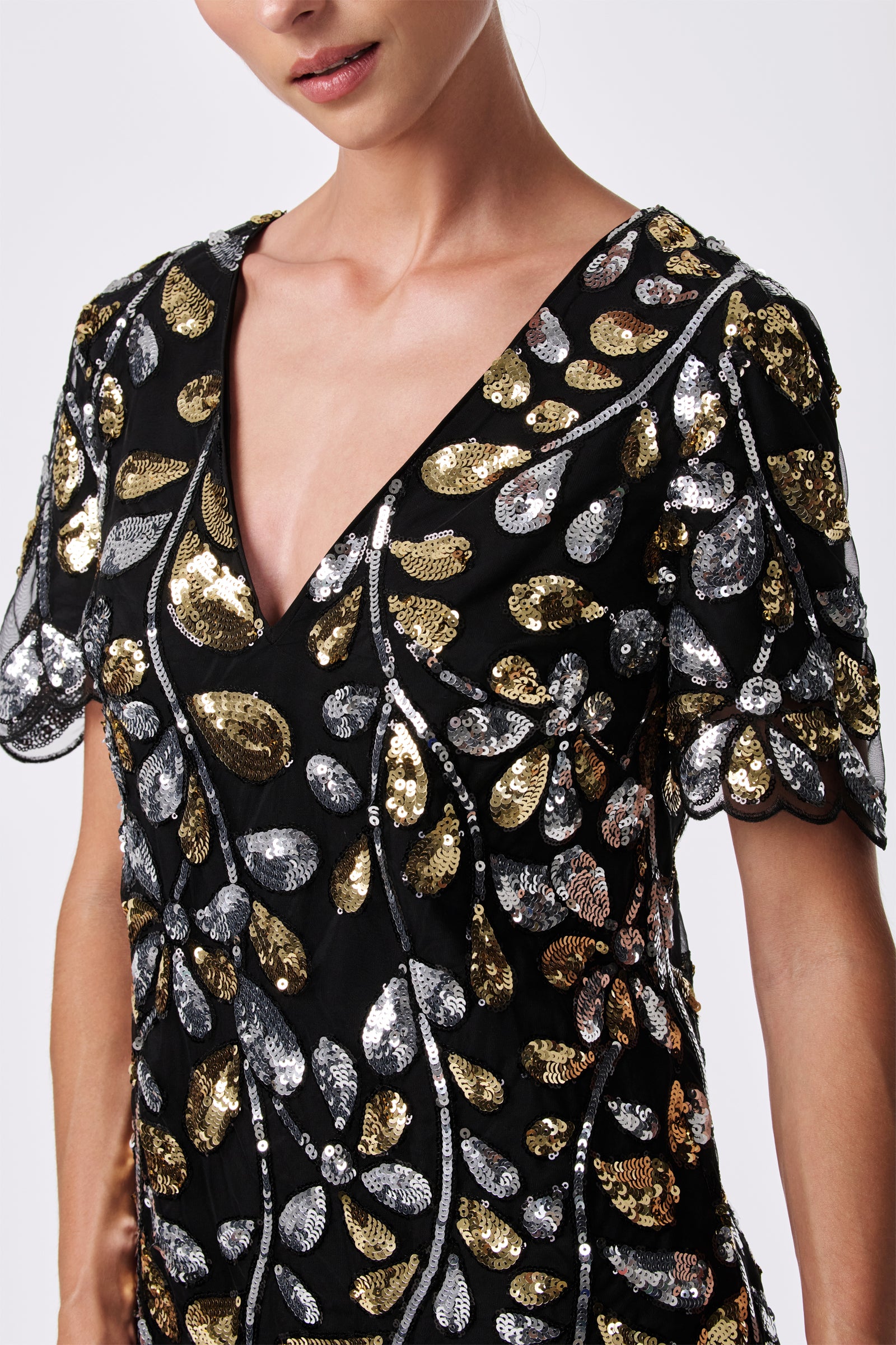 A woman wears the OBELLE 2 DRESS, a black 100% nylon piece with a deep V-neck, short sleeves, and gold and silver sequin leaf patterns. The image is cropped at her upper torso and doesn’t show her full face.