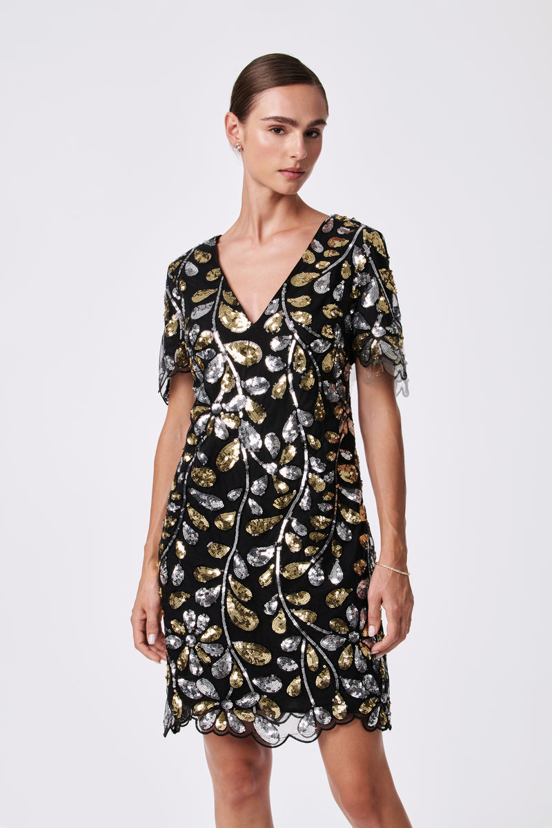 A woman stands against a plain light background wearing the OBELLE 2 DRESS, a black V-neck, short-sleeve dress made from 100% nylon with gold and silver sequin leaf patterns and a scalloped hem.