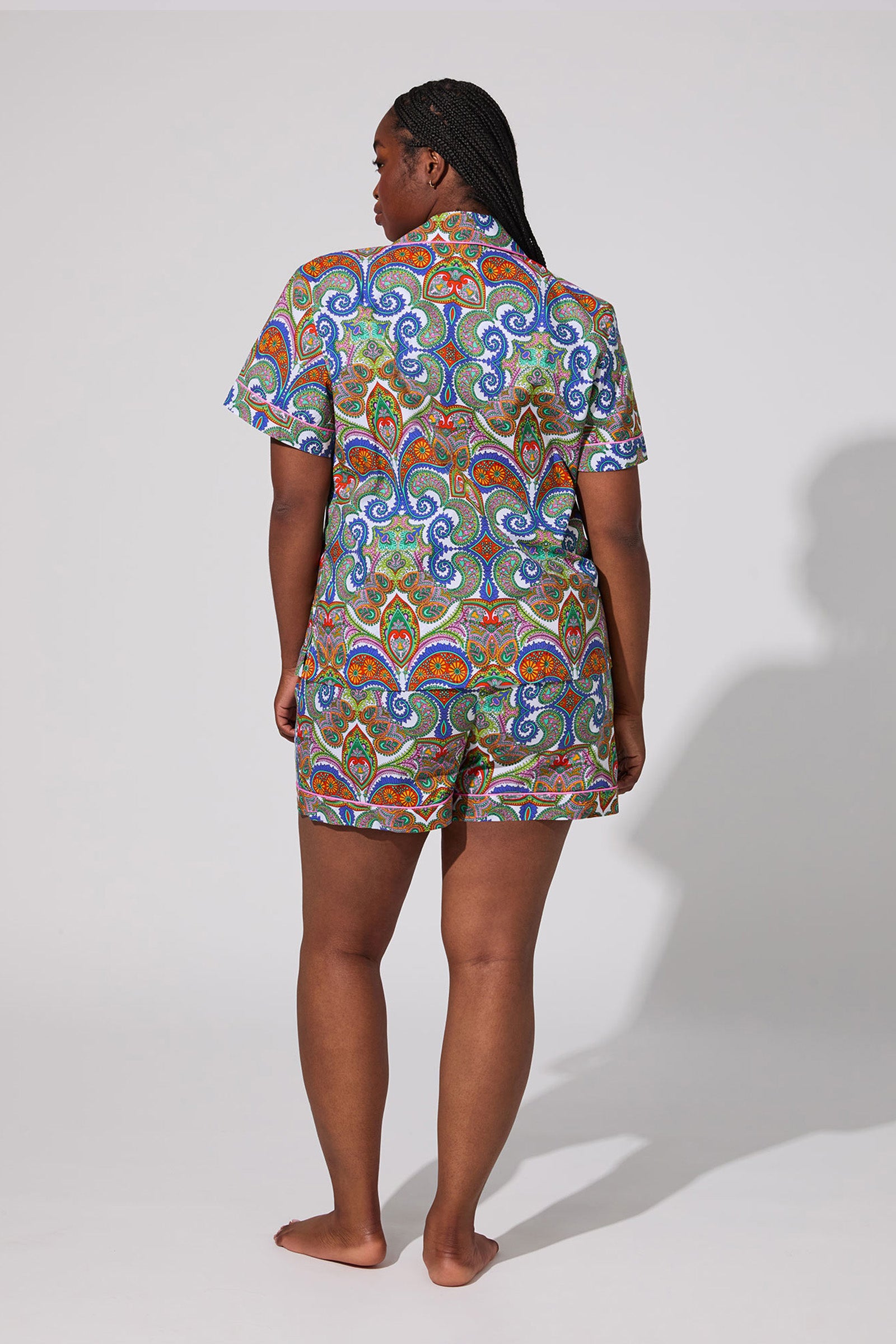 A person with long braids stands barefoot facing away, wearing the Shorty PJ Set - Paisley, a colorful short-sleeve matching set with an ornate paisley pattern, against a plain light background.