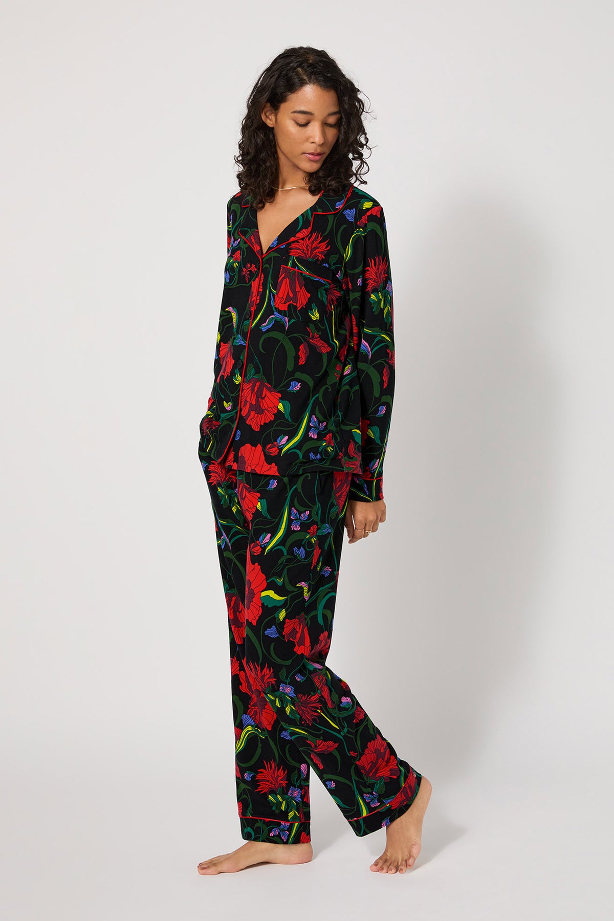 A woman with curly hair wears the Haute Floral L/S Classic Pj, a matching set with a vibrant red, purple, and green floral print on black. She stands barefoot against a plain white background, gazing down and to the side.