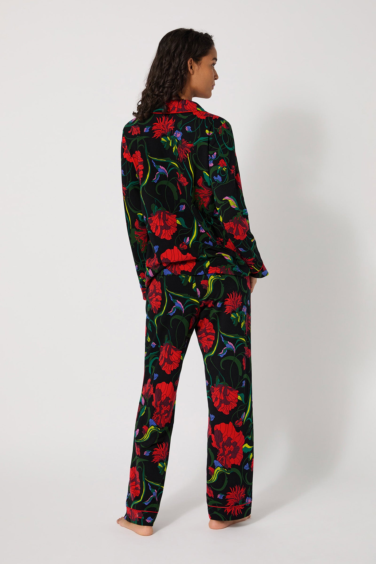 A woman stands barefoot, facing away, wearing the Haute Floral L/S Classic Pj—a matching set with a black background and bold red floral print. The long-sleeve top and loose pants are shown against a plain white background.