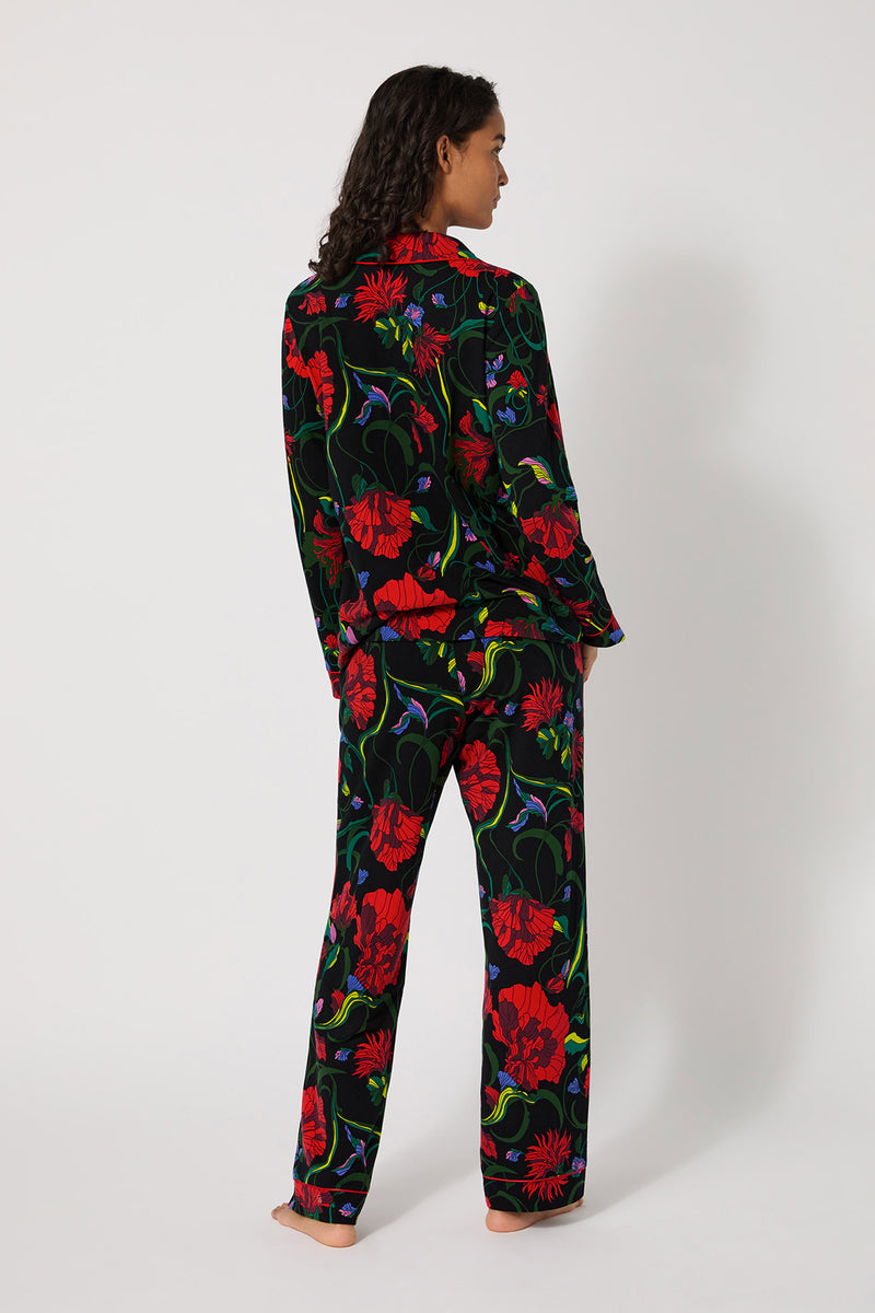 A woman stands barefoot, facing away, wearing the Haute Floral L/S Classic Pj—a matching set with a black background and bold red floral print. The long-sleeve top and loose pants are shown against a plain white background.