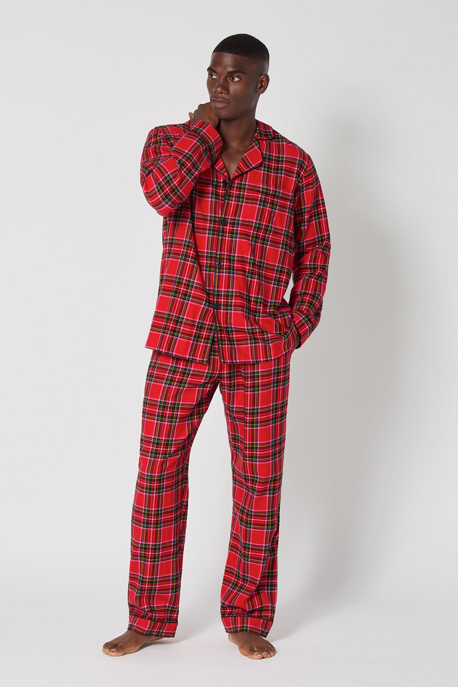 A man stands barefoot against a plain background, wearing the Christmas Plaid Men Classic Pj set with long sleeves and long pants. He looks to the side, touching his face with one hand.
