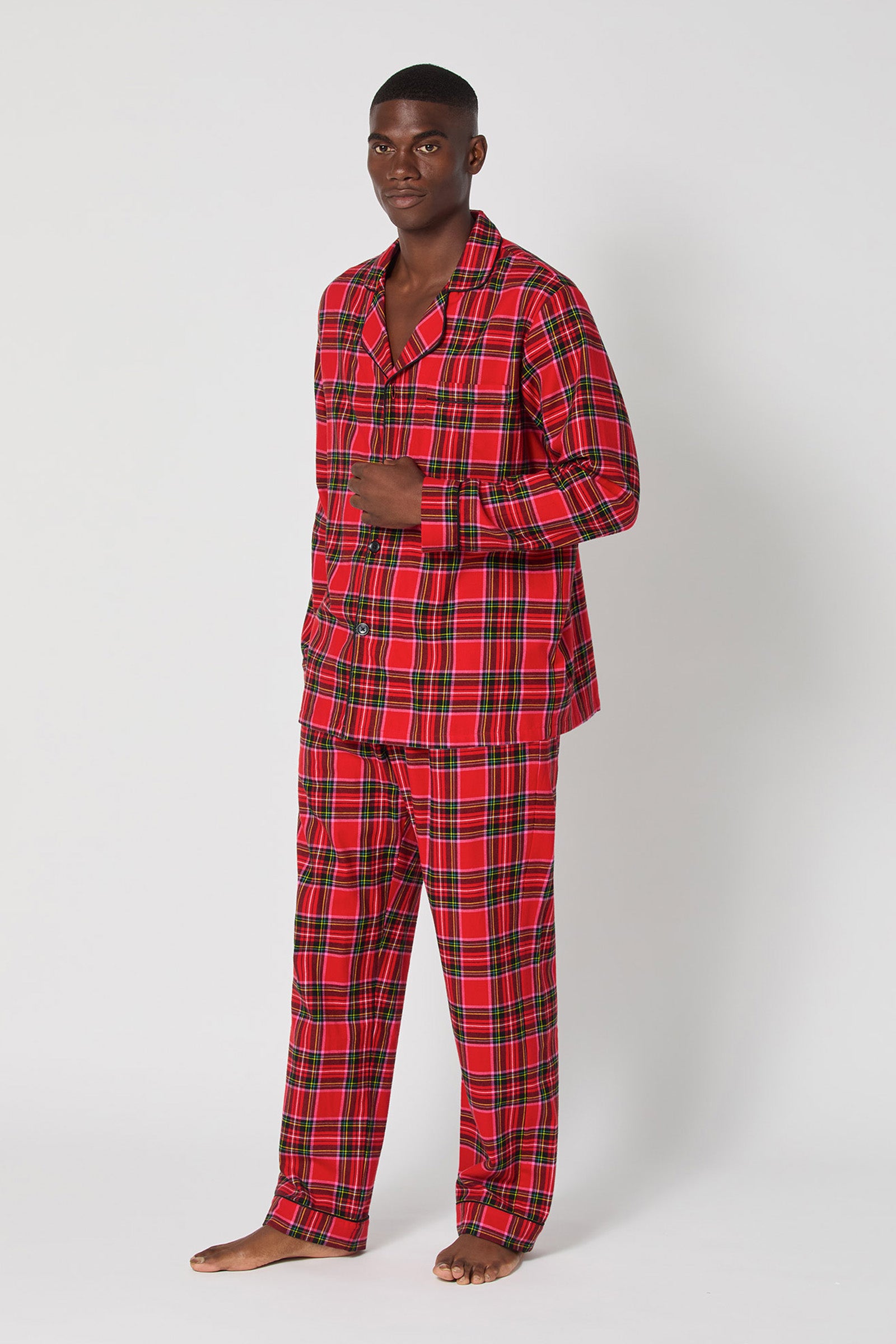 A man stands barefoot against a plain white background, wearing the Christmas Plaid Men Classic Pj set featuring a matching red and black plaid button-up shirt and long pants.