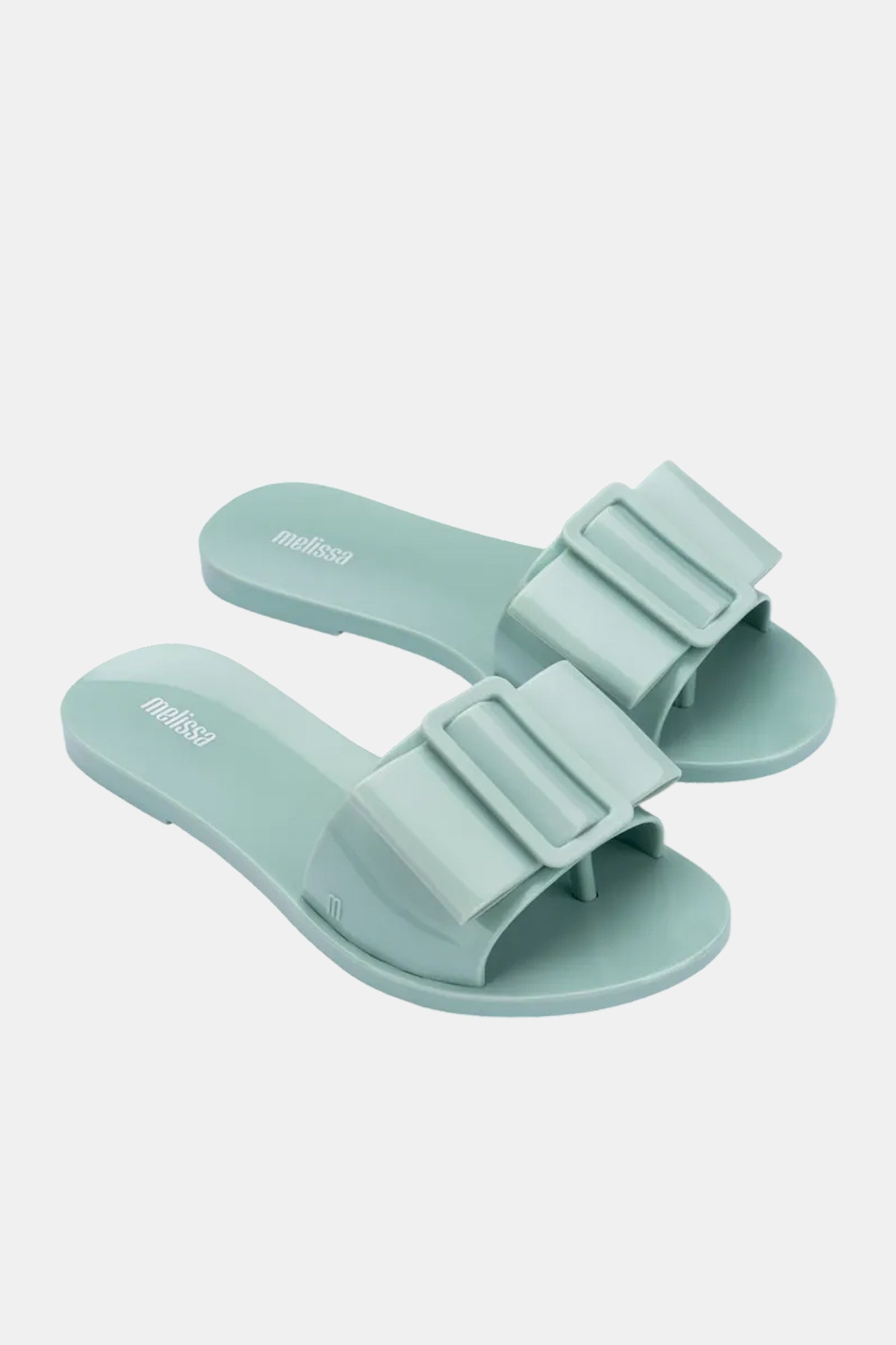 The Melissa Babe Sandal is a pair of light mint green slides with wide straps, oversized buckle details, a glossy finish, and a simple modern design.