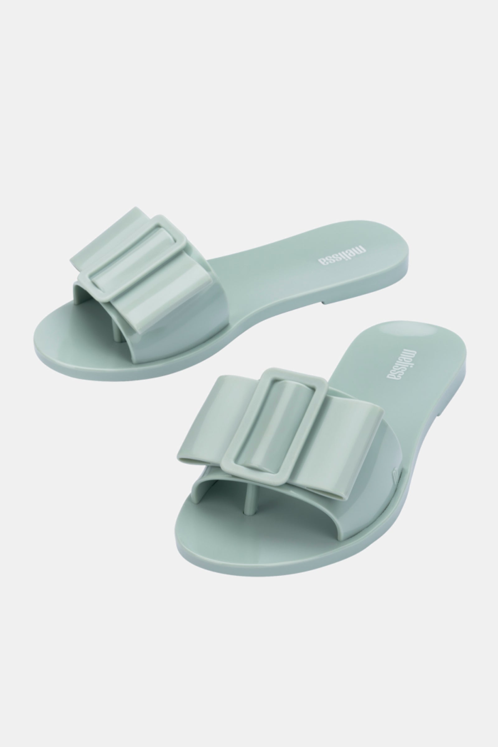 The Melissa Babe Sandal is a pair of light green slide sandals featuring a wide, sculpted bow design on the strap, displayed against a plain white background.