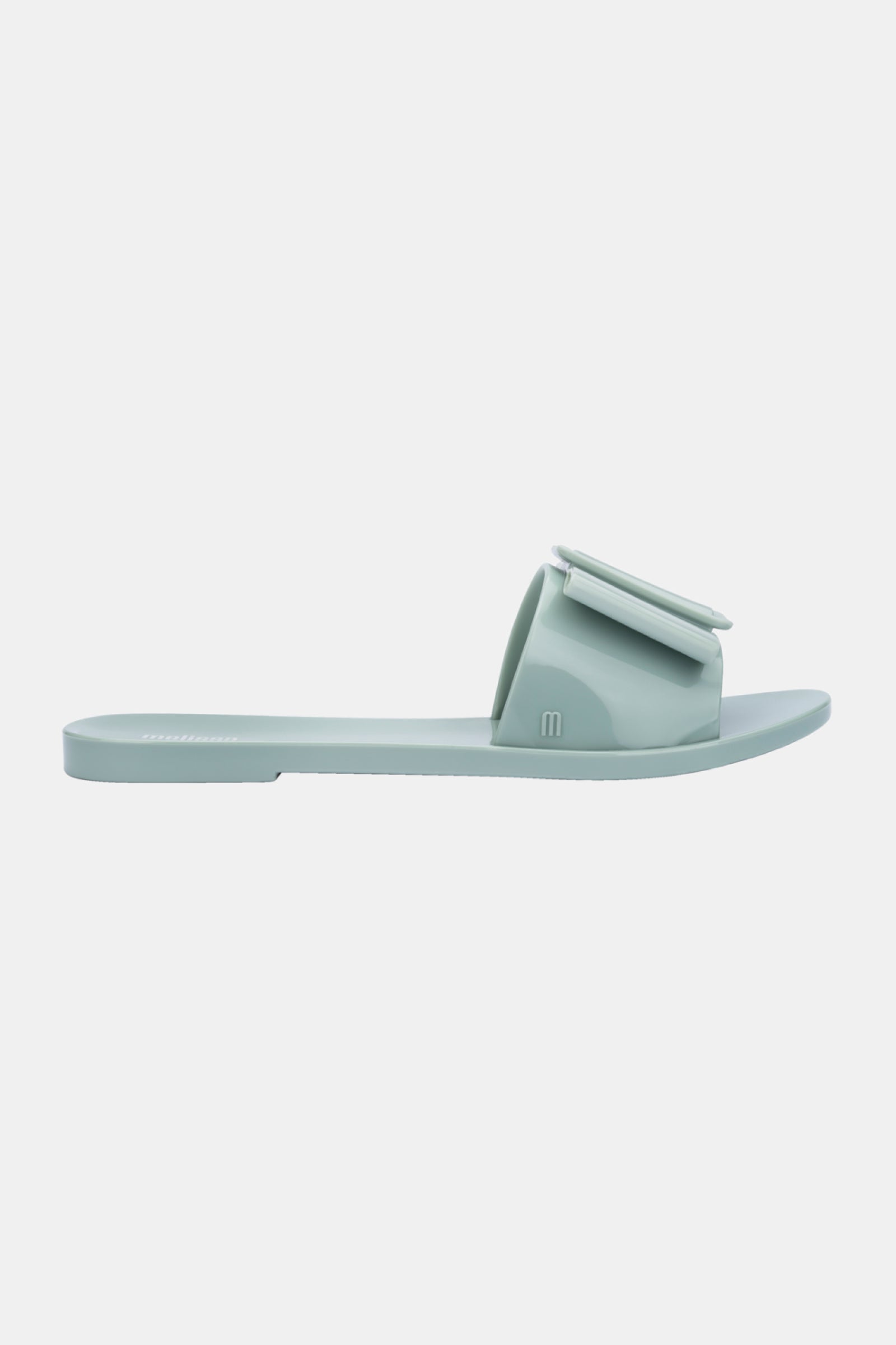 The Melissa Babe Sandal is a minimalist, light blue slide with a flat sole and wide strap topped by a large decorative bow, shown in side profile against a plain white background.