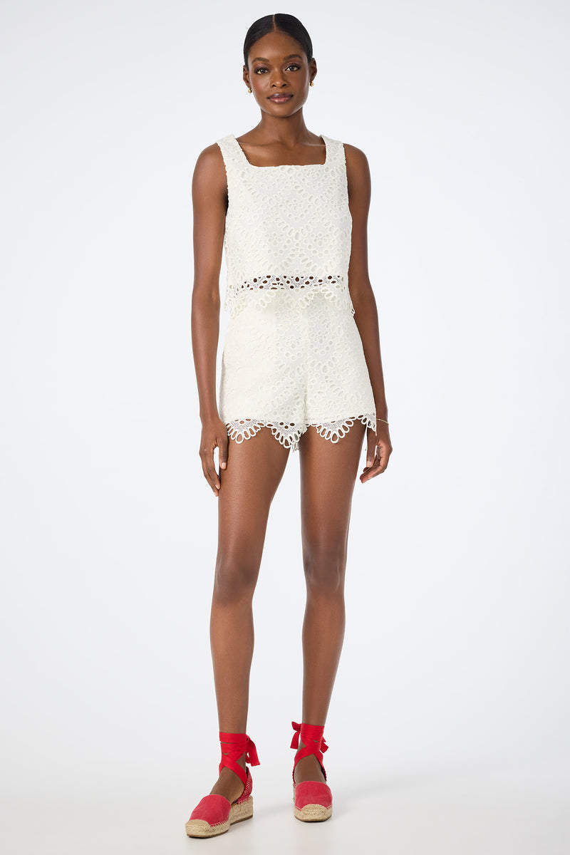 A woman wears the Albi Lace Crop Top—sleeveless, white, and cropped—paired with matching high-waisted shorts with scalloped edges. She completes the look with red espadrille ankle-tie shoes against a plain white backdrop.