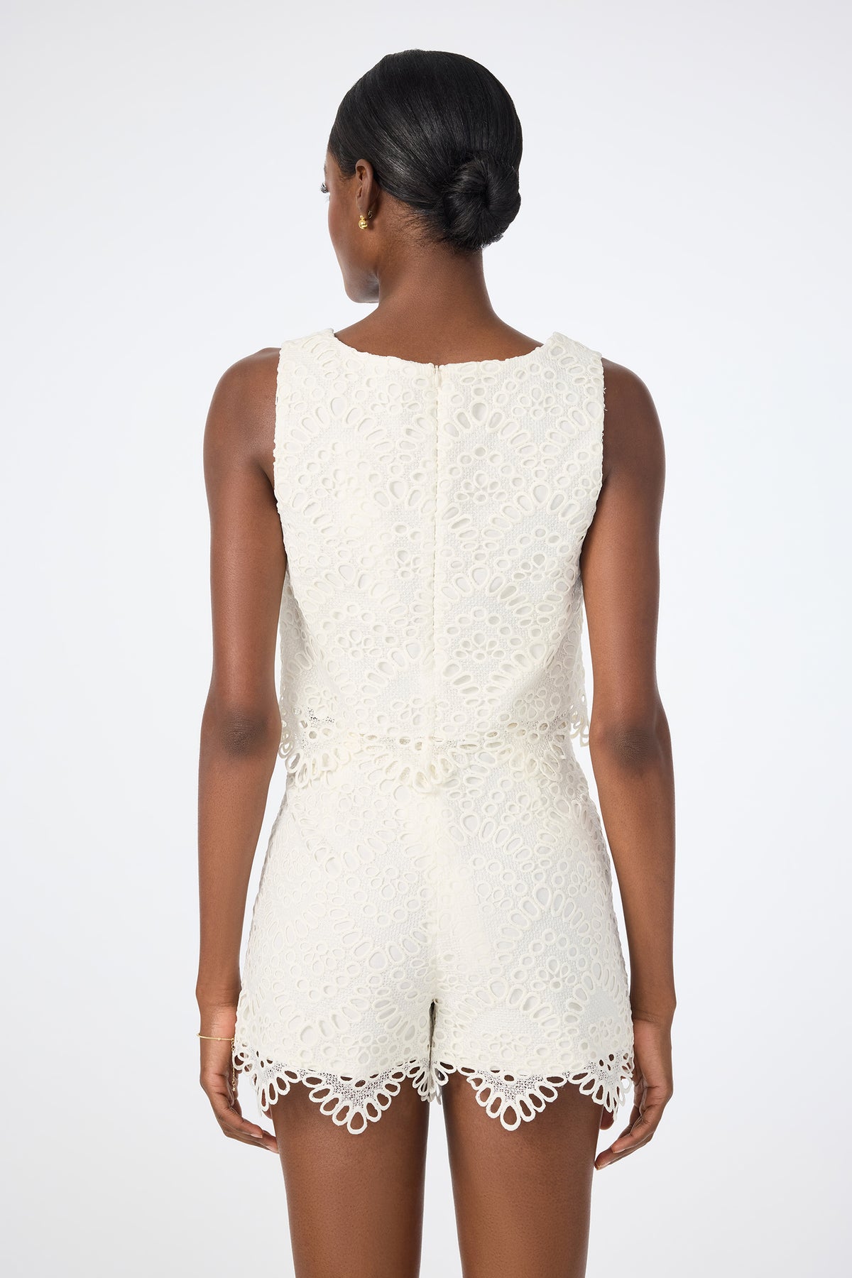 A woman with dark hair in a low bun is seen from behind, wearing the Albi Lace Crop Top—a sleeveless, white lace top with scalloped edges and intricate cut-out patterns—against a plain light background.