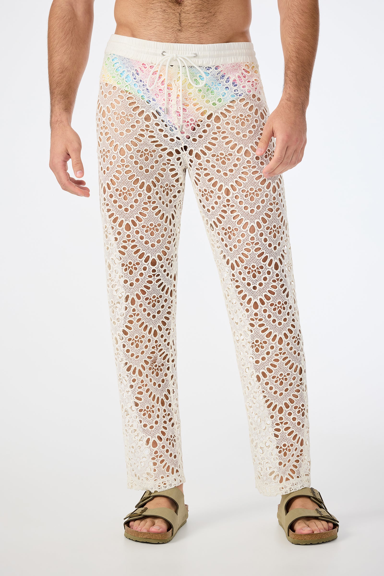 A person wears the Venice Beach Resort Pant - Lerins Lace in Whitewash, featuring a see-through crocheted lace design and drawstring waist. Styled with brown sandals, the detailed pattern hints at colorful underwear underneath.