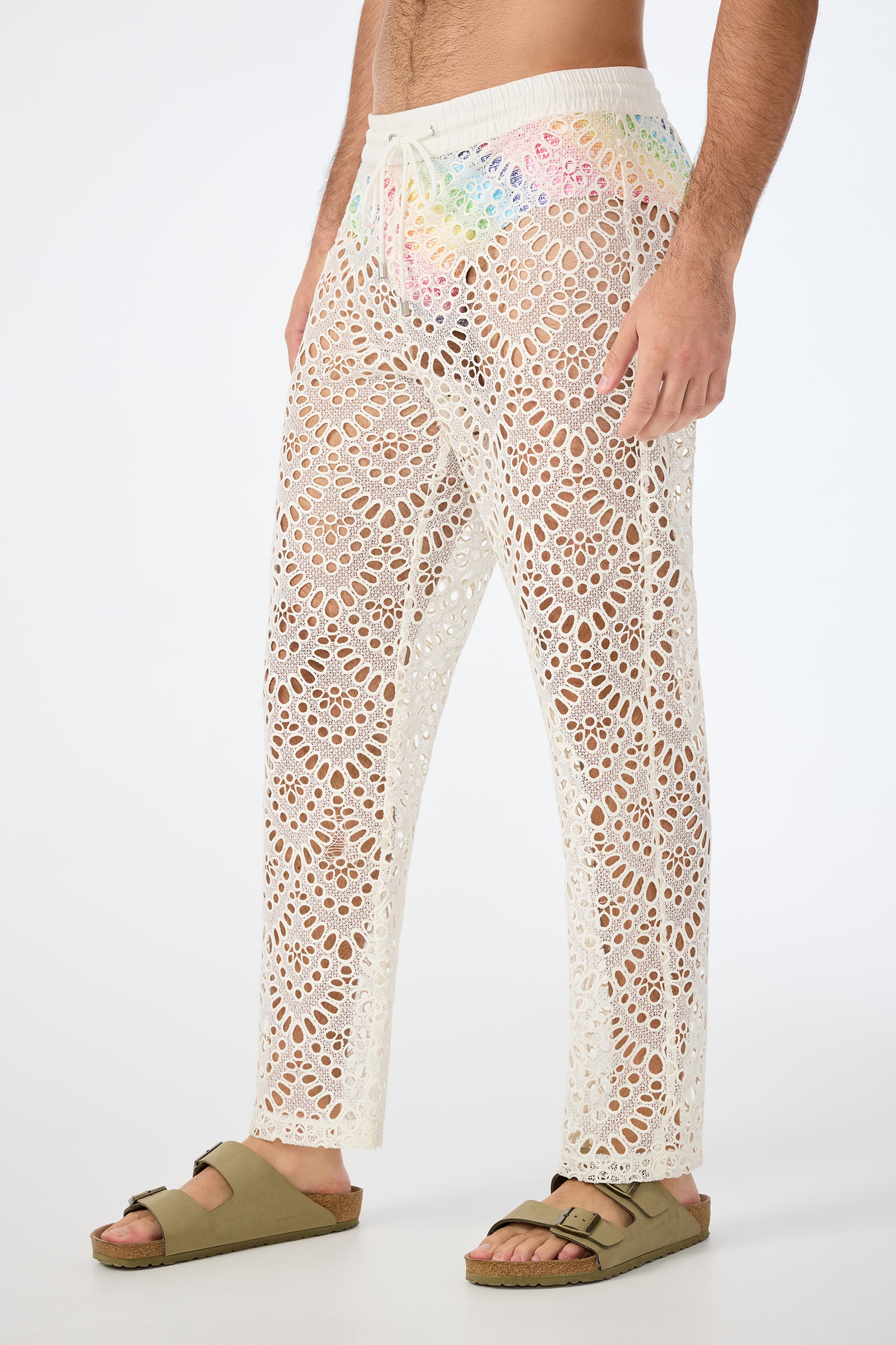 A person wears the Venice Beach Resort Pant - Lerins Lace, featuring white, see-through floral lace over rainbow underwear and beige sandals, standing against a white background.