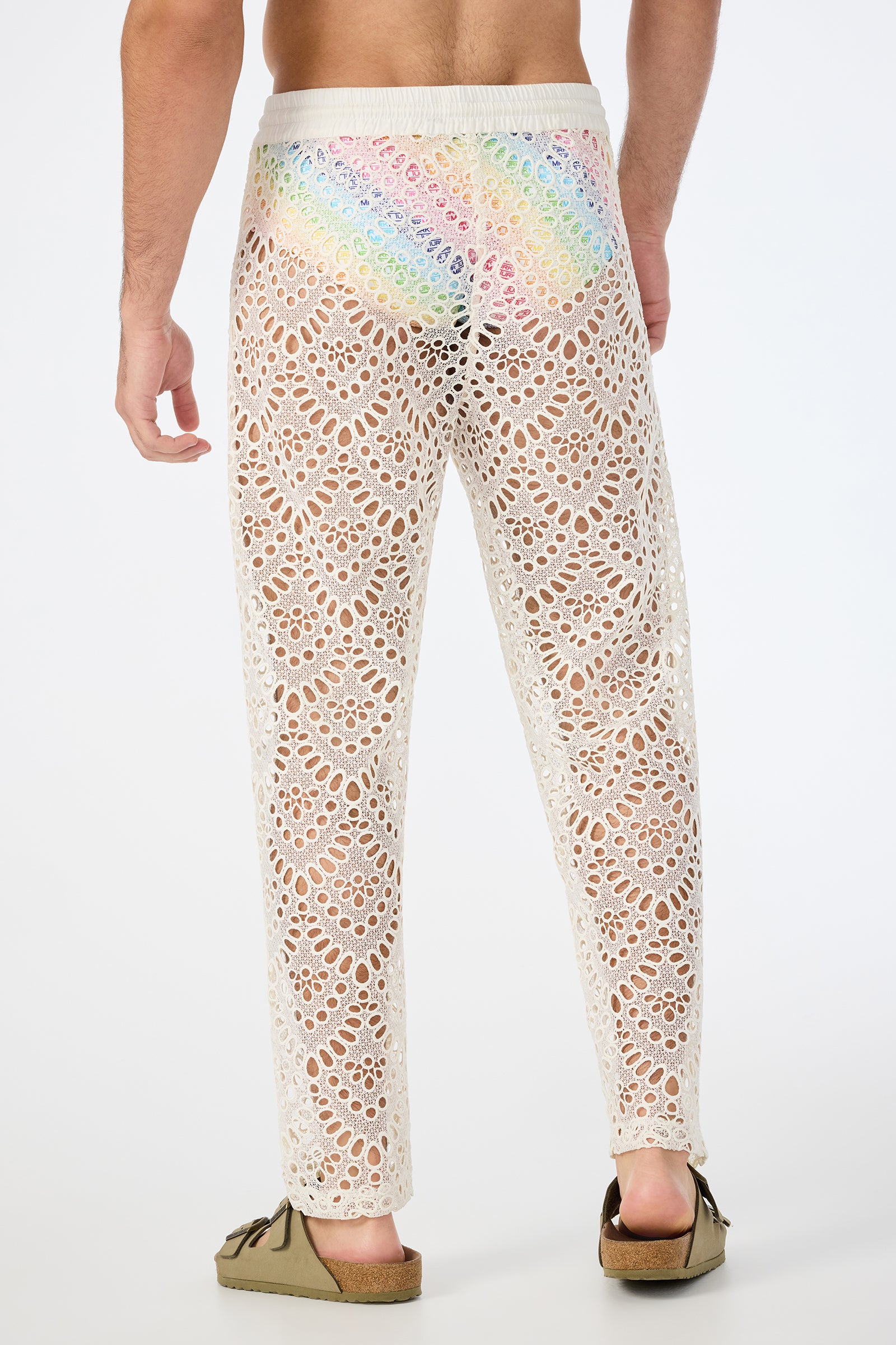 A person stands facing away, wearing the Venice Beach Resort Pant - Lerins Lace in cream over rainbow briefs and brown slip-on sandals; the intricate lace is see-through, revealing the colorful underwear beneath.