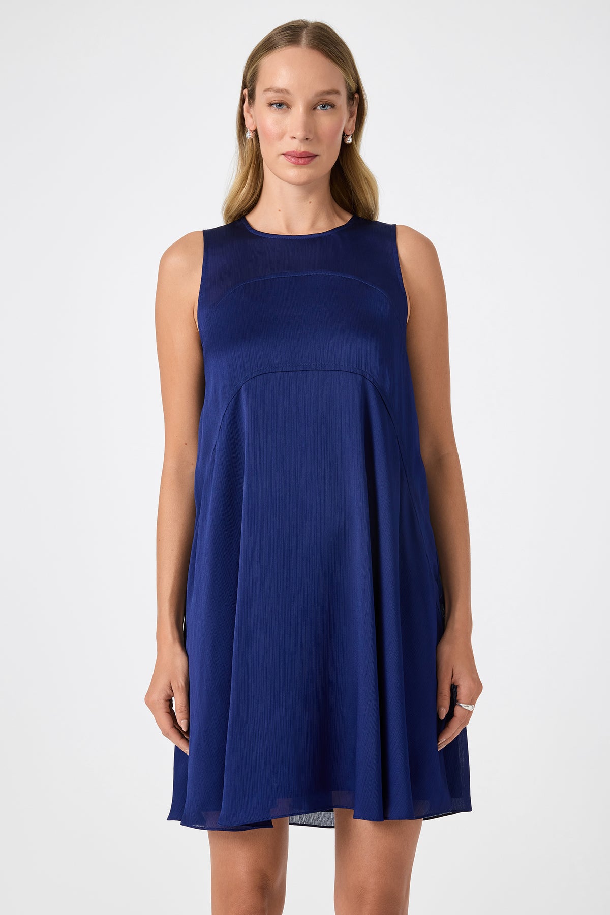 A woman with long blonde hair wears the Mauvie Sleeveless Mini Dress, a dark blue knee-length piece. She stands facing forward against a plain white background with a neutral expression.