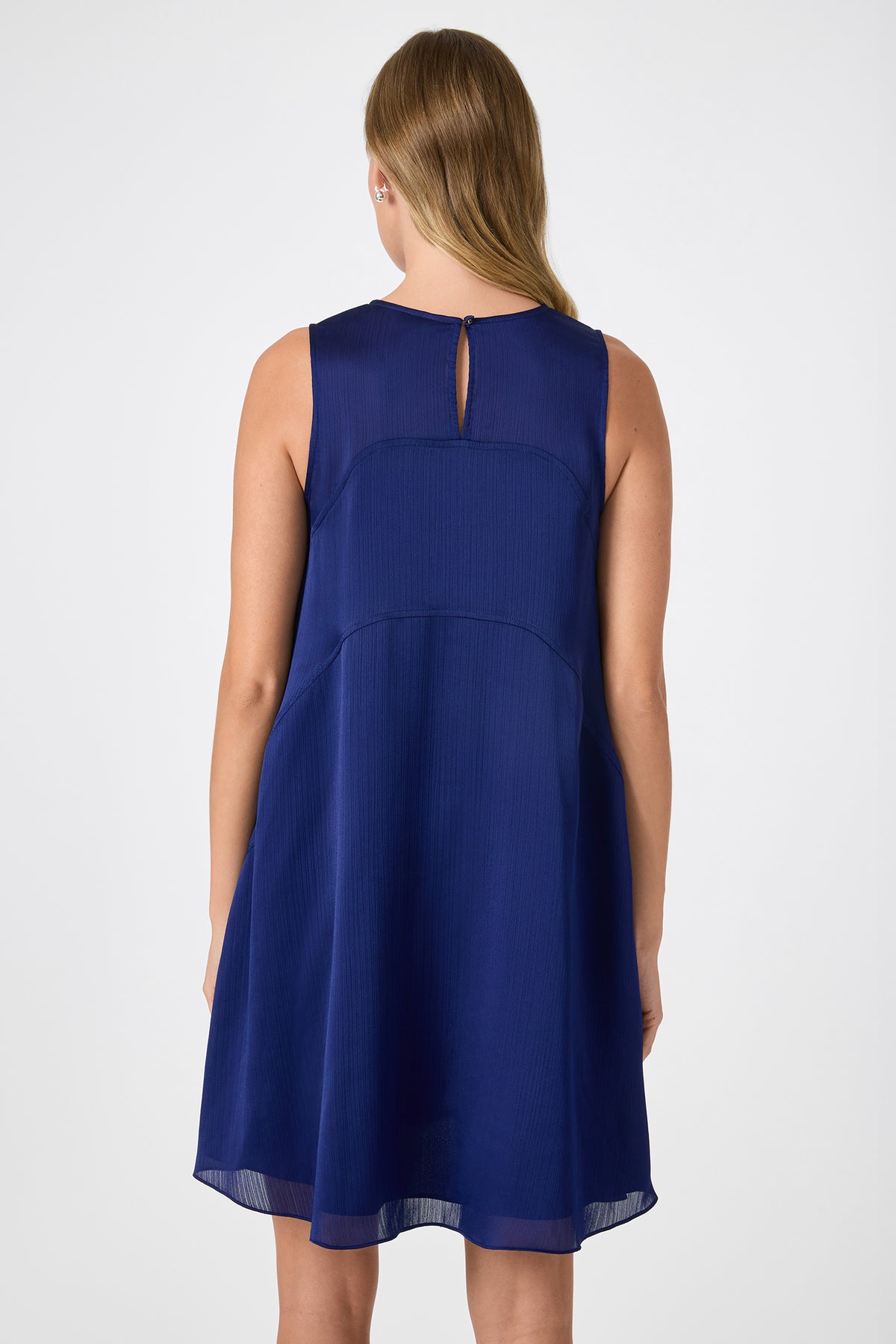 A woman with light brown hair stands with her back to the camera, wearing a Mauvie Sleeveless Mini Dress in dark blue against a plain, light background.