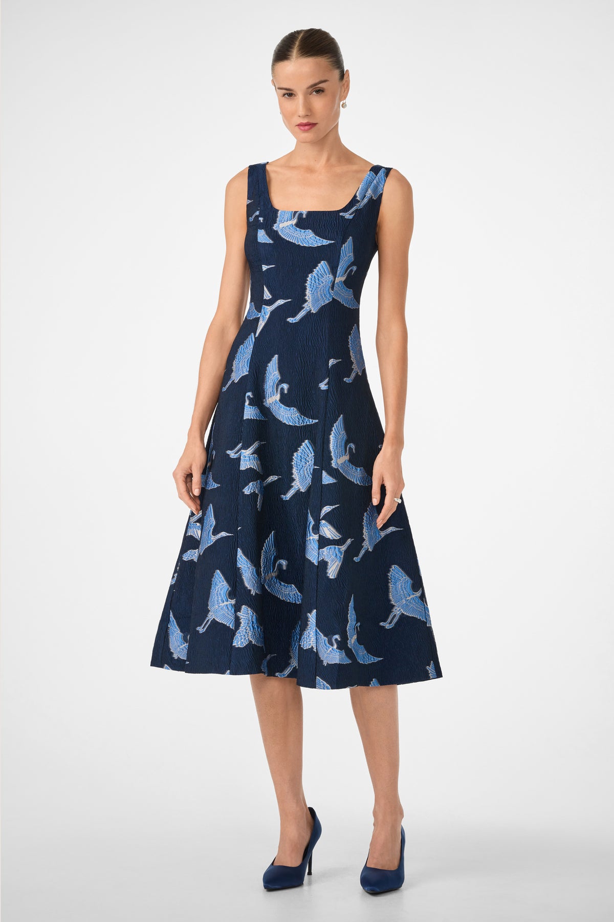 A woman with her hair tied back and a neutral expression stands against a white background wearing the Valor Sleeveless Midi Dress in Sumi Blue Tsuru, styled with matching blue high-heeled shoes.