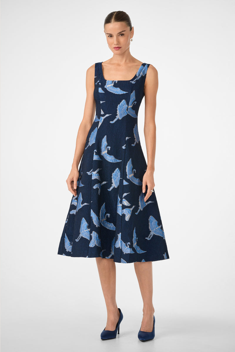 A woman with her hair tied back and a neutral expression stands against a white background wearing the Valor Sleeveless Midi Dress in Sumi Blue Tsuru, styled with matching blue high-heeled shoes.