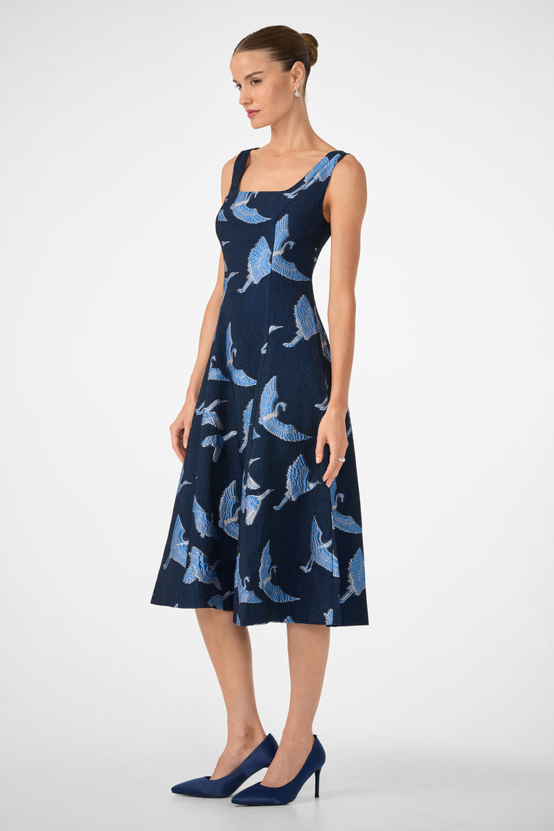 A woman in the Valor Sleeveless Midi Dress, a navy blue knee-length style with a blue bird pattern, stands in profile on matching heels. Her hair is in a neat bun against a plain white background.
