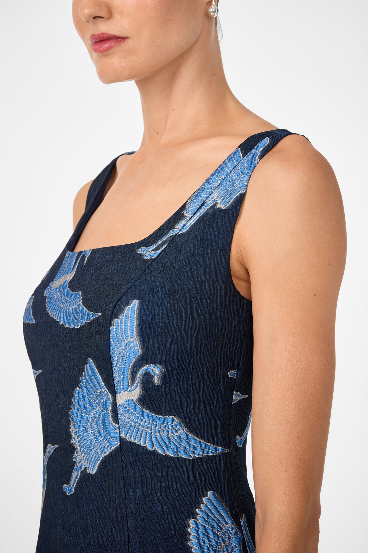 A woman is shown from shoulders to above the waist wearing the Valor Sleeveless Midi Dress, featuring a textured dark blue fabric with a blue crane bird pattern, set against a plain background.