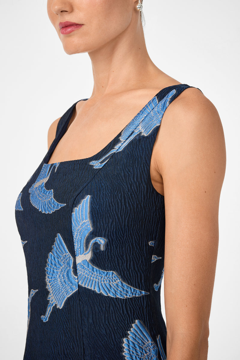 A woman is shown from shoulders to above the waist wearing the Valor Sleeveless Midi Dress, featuring a textured dark blue fabric with a blue crane bird pattern, set against a plain background.