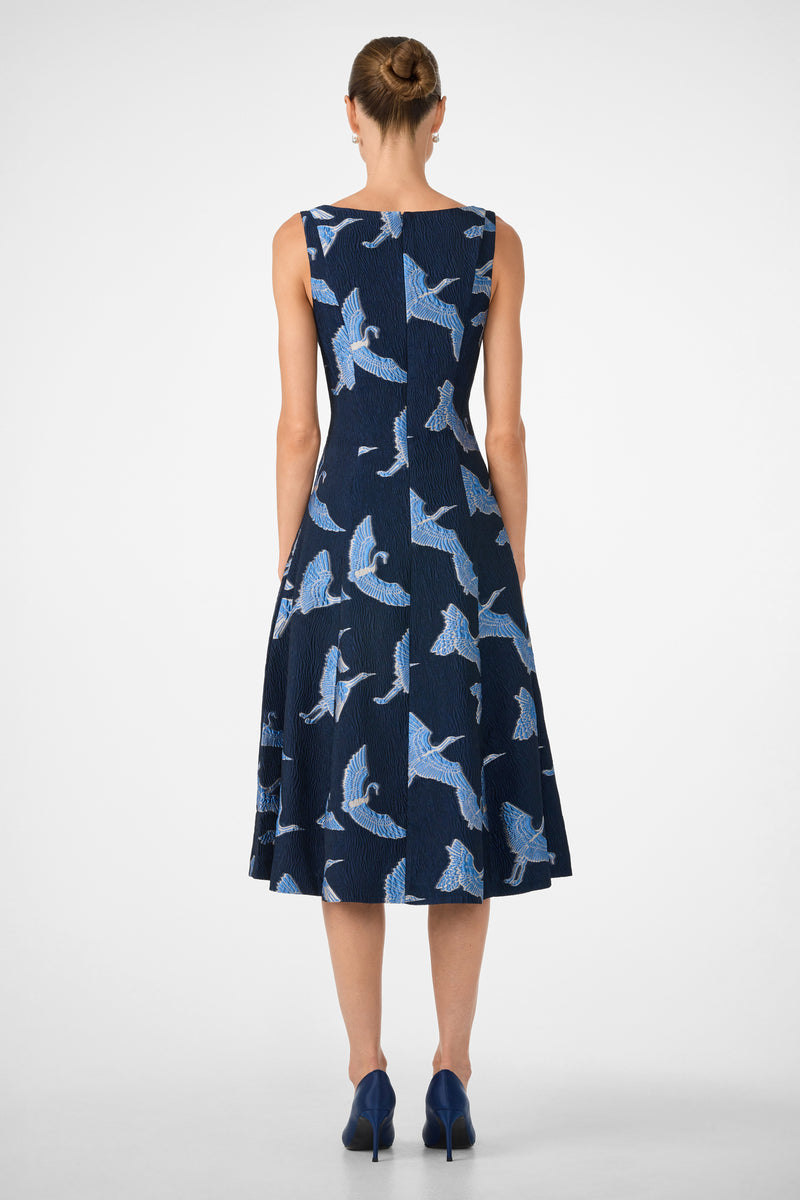 A woman with her hair in a bun models the Valor Sleeveless Midi Dress, featuring a navy blue bird print, and blue heels, standing with her back to the camera against a plain white background.