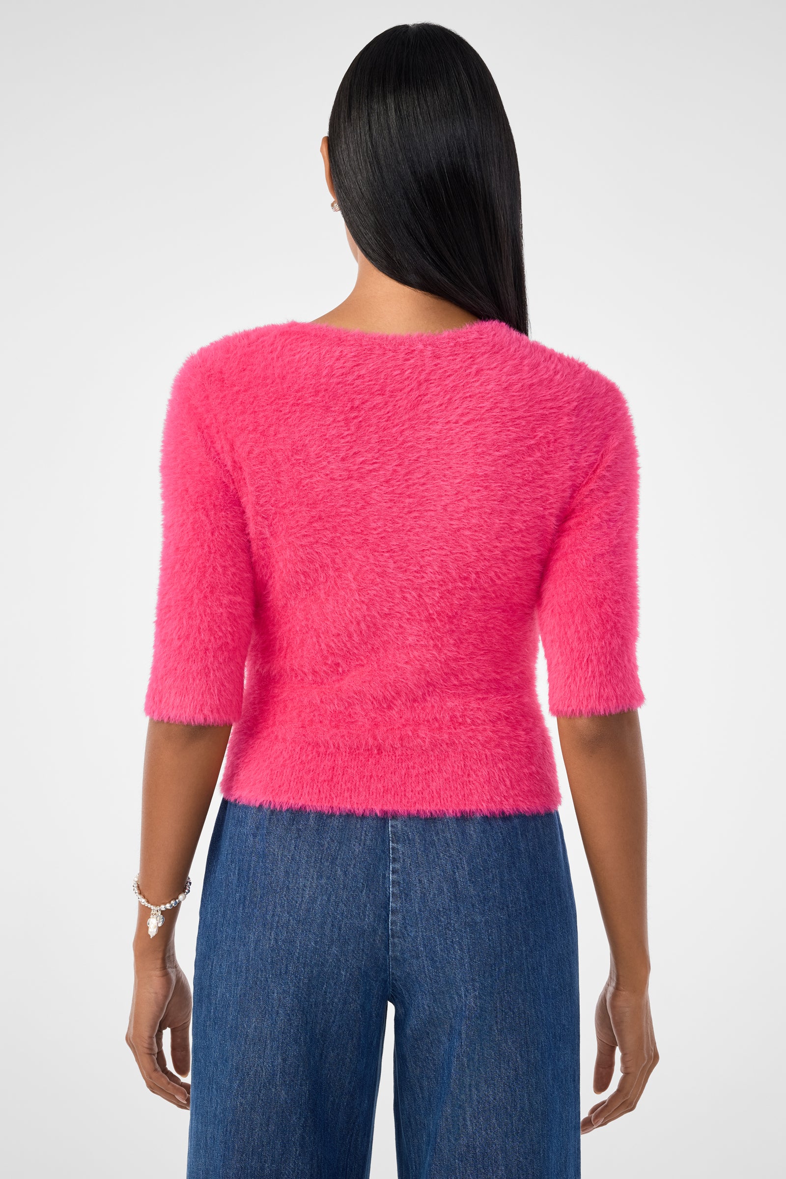 A woman with straight, dark hair wears the Maliah Fuzzy Sweater in bright pink and blue jeans. She faces away from the camera, showing a bracelet on her left wrist against a plain white background.